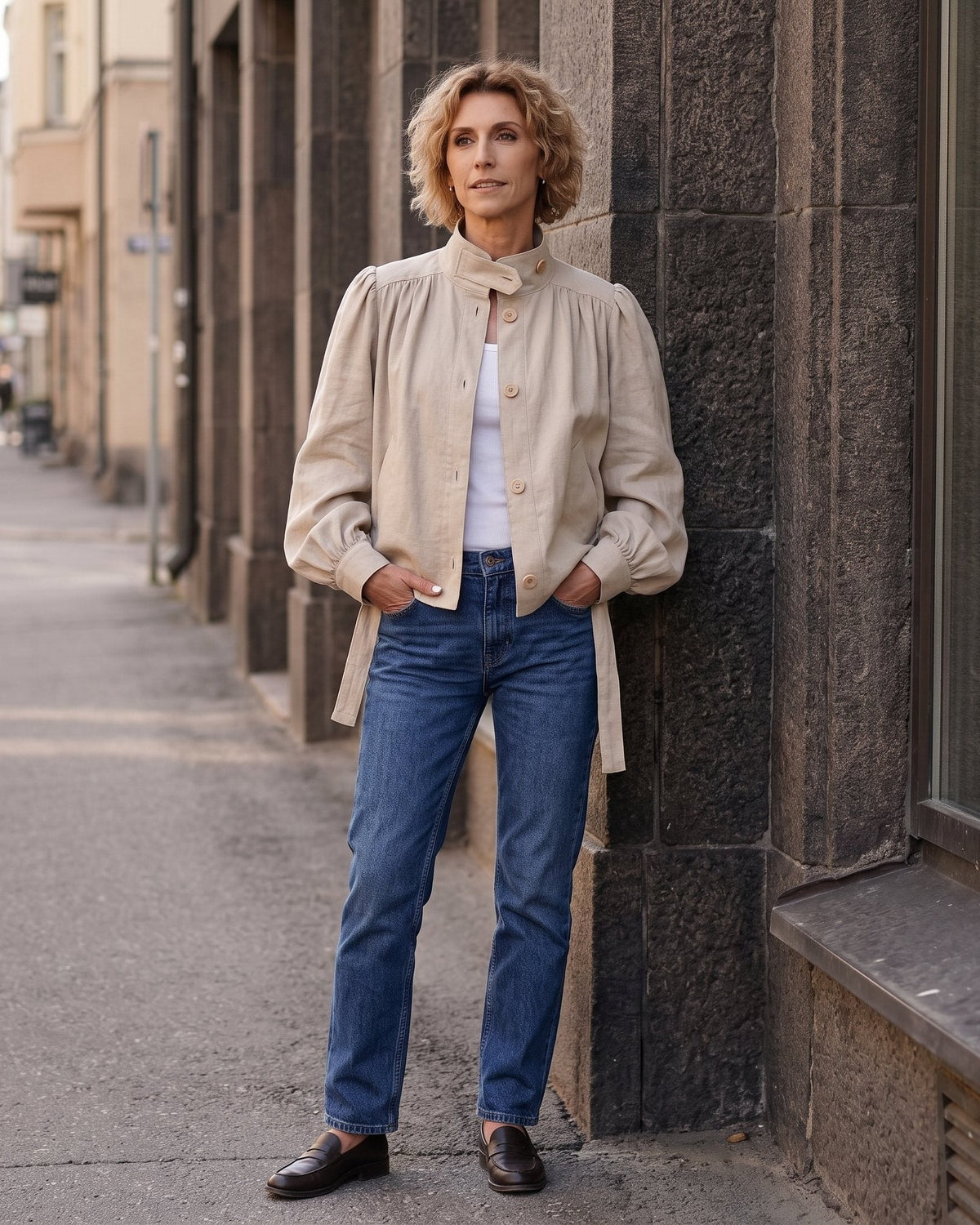 A woman stands on a city sidewalk leaning against a dark stone building, wearing a beige high-neck jacket over a white top with blue jeans and dark brown loafers. #Color_Beige
