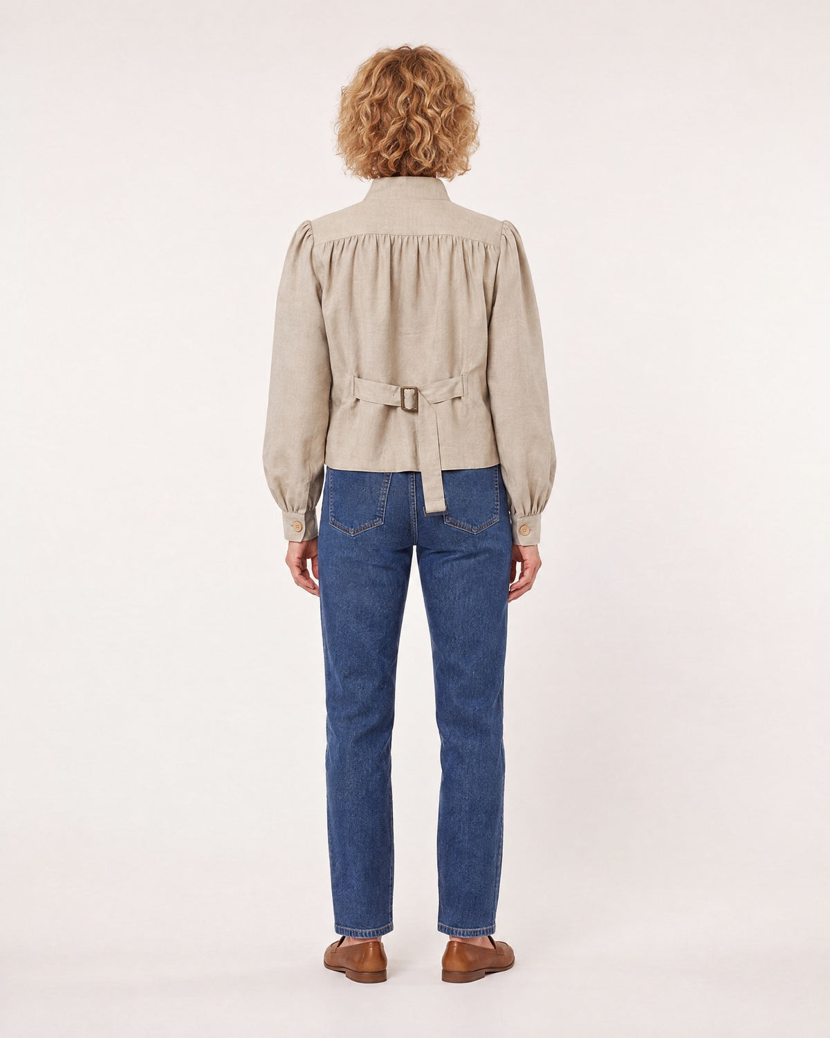 A woman stands with her back to the camera against a plain light studio background, wearing a light beige linen jacket with a stand collar, gathered shoulders, and a tie belt with buckle at the back, paired with blue jeans and brown loafers. #Color_Beige