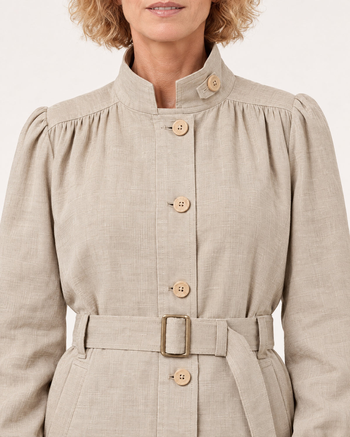 Close-up of a light beige linen jacket showing the stand collar with tab-button closure, wooden buttons, and tie belt with buckle. #Color_Beige