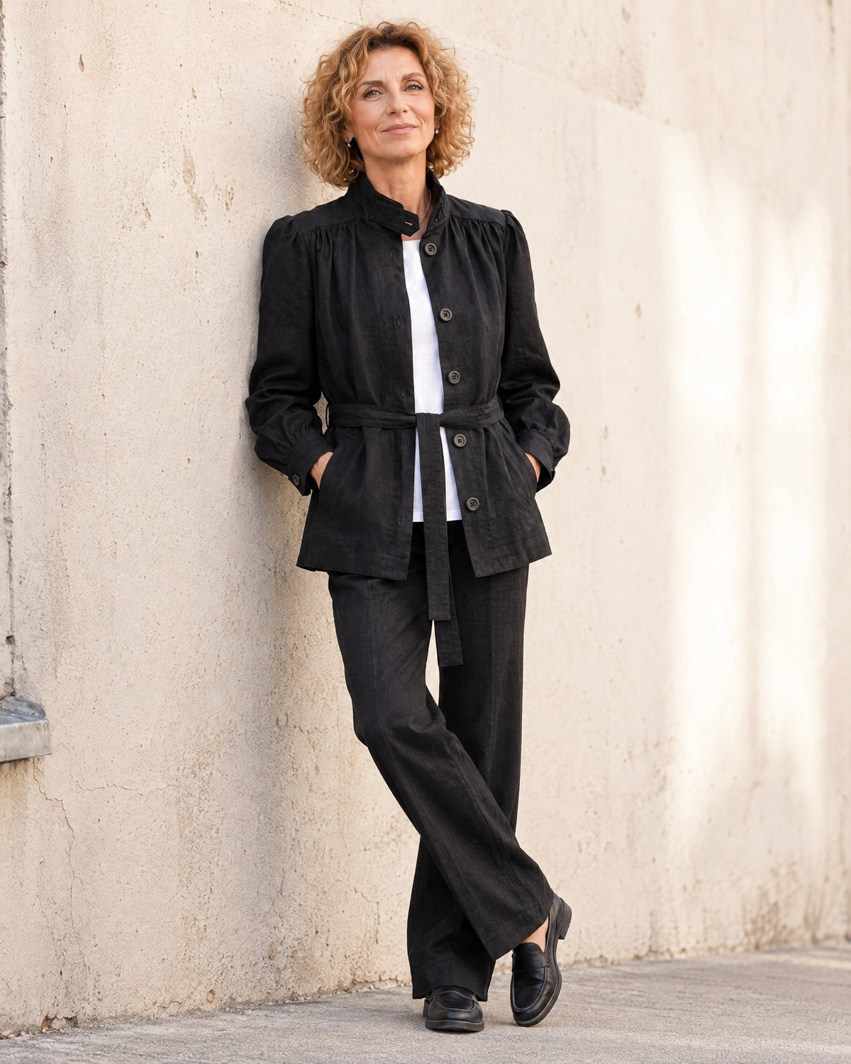 A woman stands full length against a light concrete wall, wearing a black linen jacket with a stand collar, gathered shoulders, and tie belt, over a white top with dark trousers and dark shoes. #Color_Black