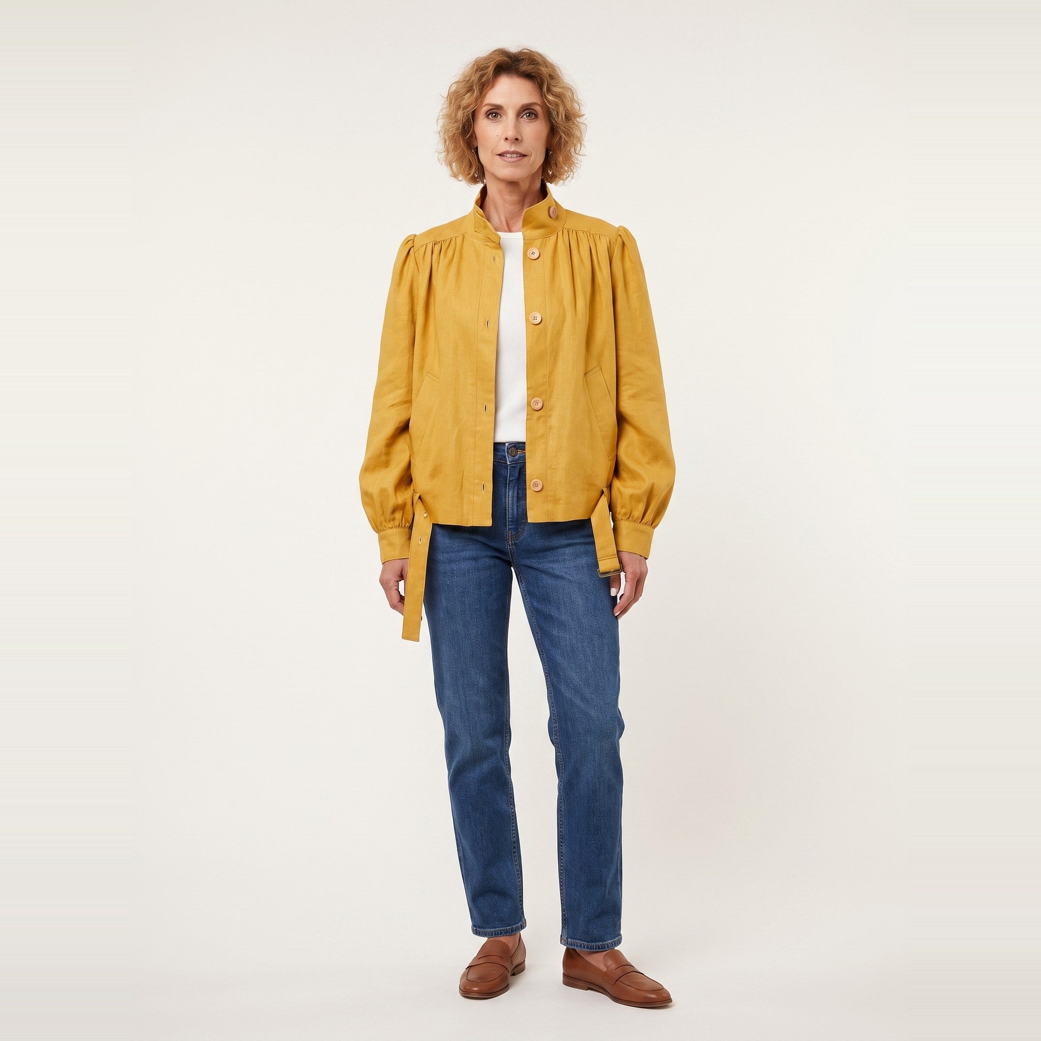 A woman stands against a plain light background wearing a mustard yellow button-front jacket with gathered sleeves, blue jeans, a white top, and brown shoes.
