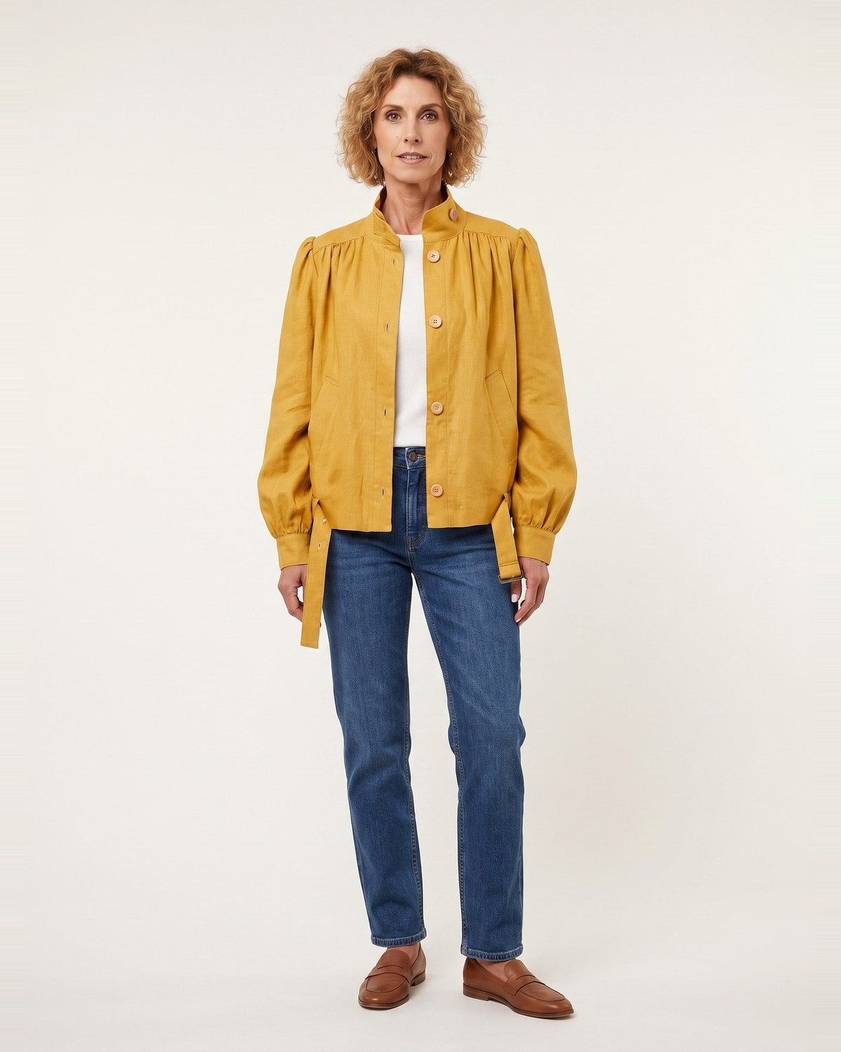 A woman stands against a plain light background wearing a mustard yellow button-front jacket with gathered sleeves, blue jeans, a white top, and brown shoes. #Color_Ochre