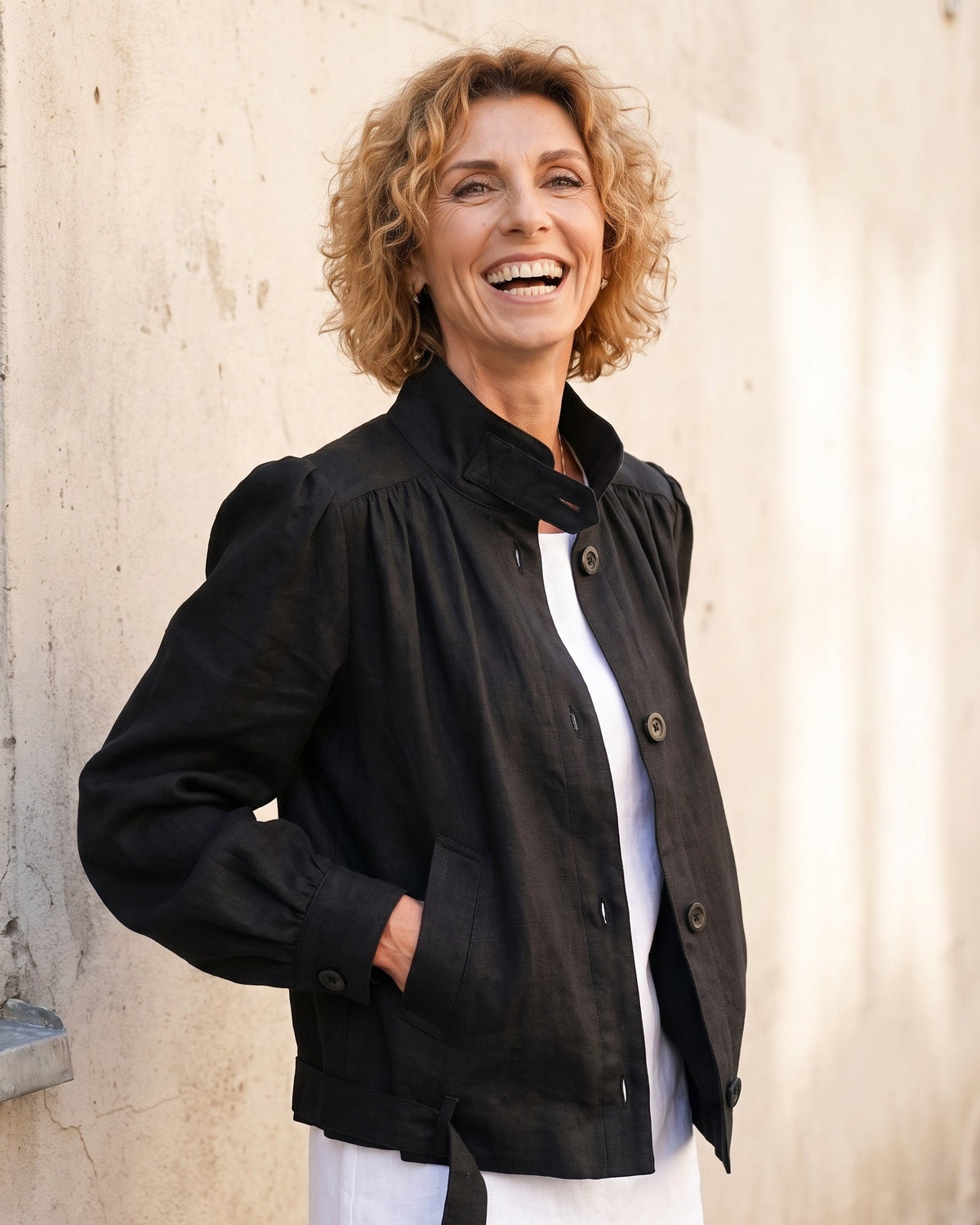 A smiling woman with curly blonde hair stands outdoors against a light beige wall, wearing a black button-front jacket over a white top. #Color_Black