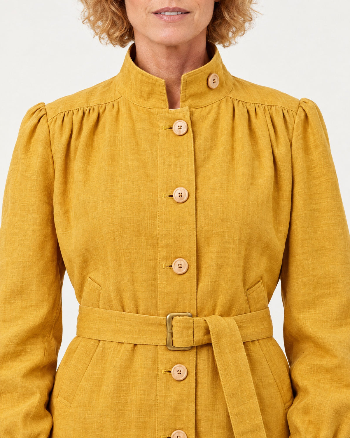 Close-up of a mustard-ochre linen jacket showing the stand collar with tab-button closure, wooden buttons, and tie belt with buckle. #Color_Ochre