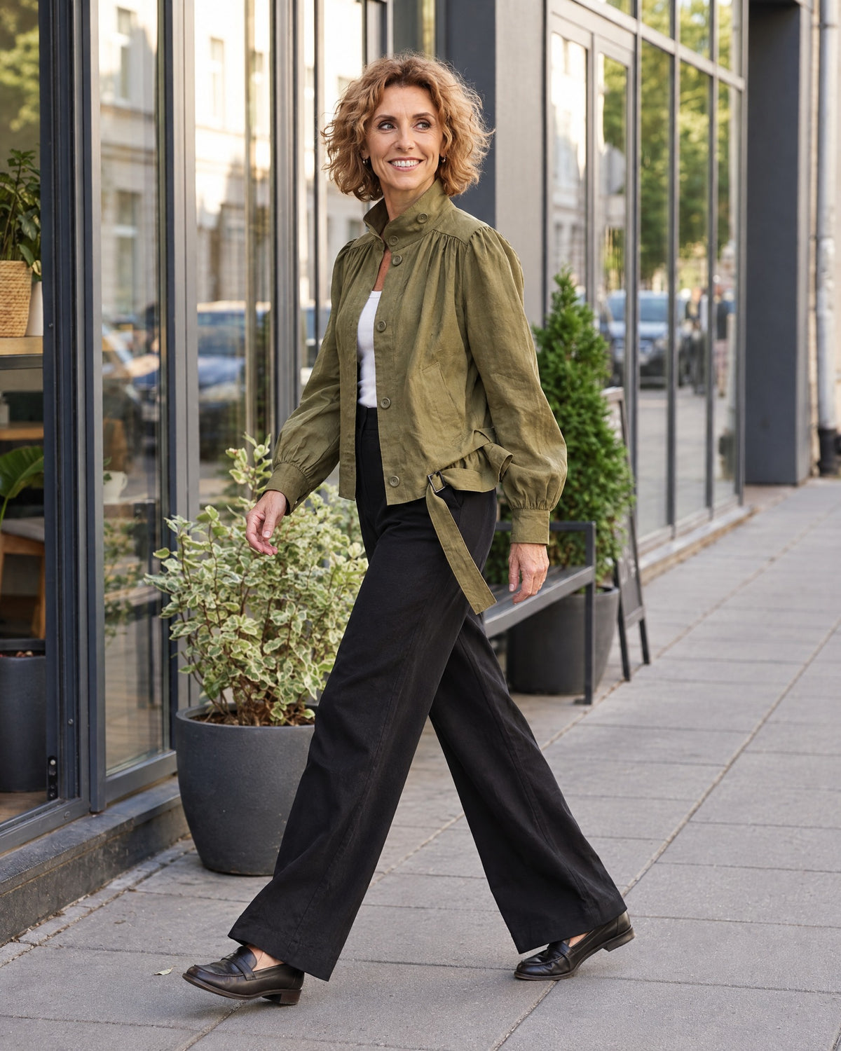 A woman walks along a city sidewalk in profile, wearing an olive green linen jacket with a stand collar and tie belt, paired with black wide-leg trousers and dark loafers. #Color_Olive green