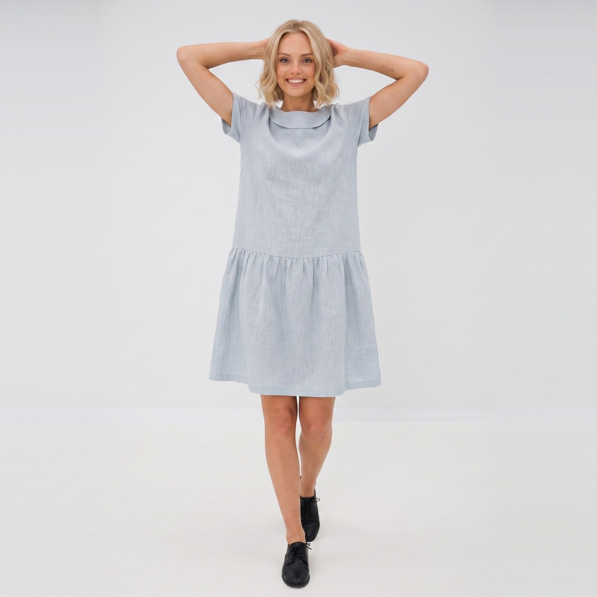 A woman modeling a light blue short-sleeve linen dress with a tiered skirt, standing against a plain white studio background.