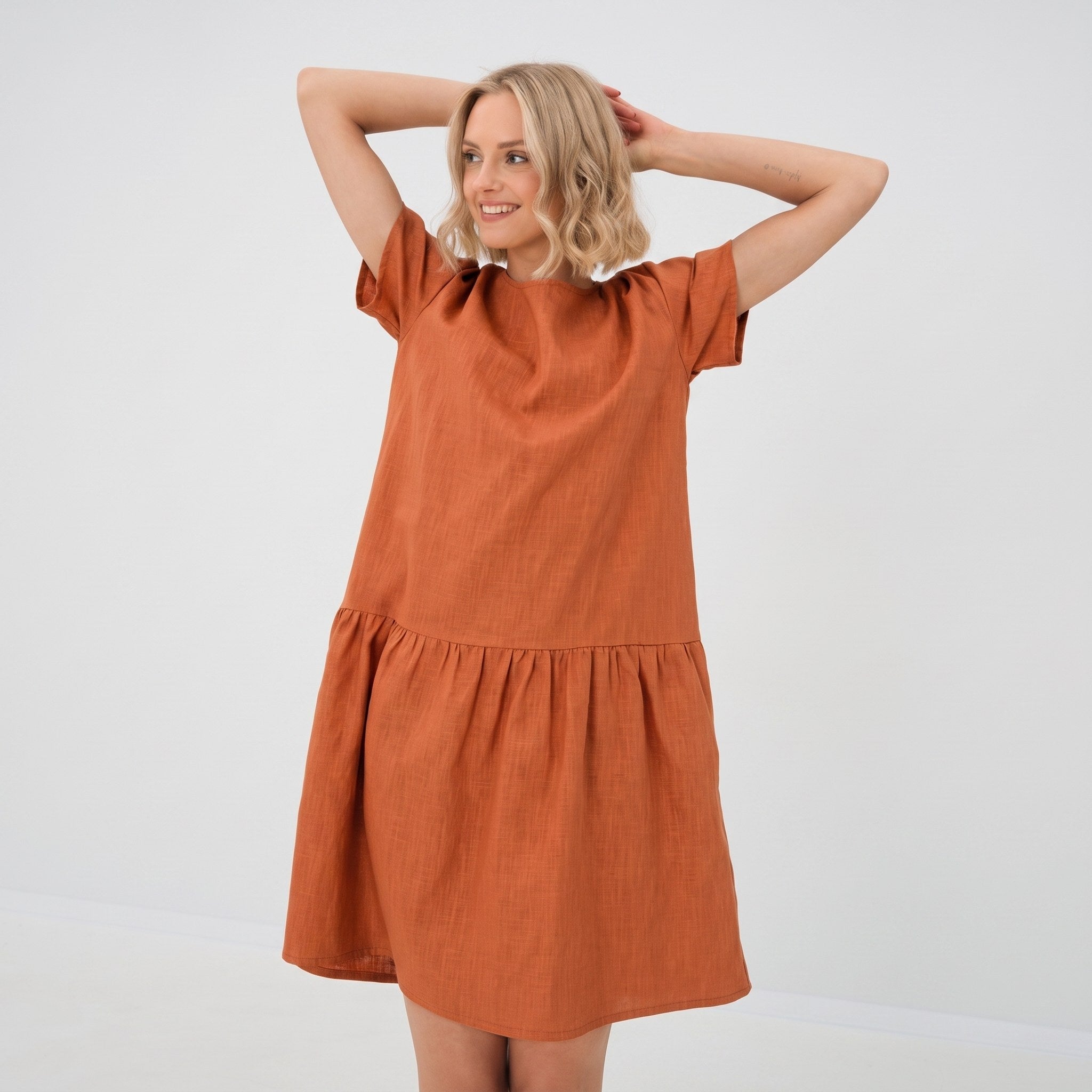 A woman modeling a short-sleeve terracotta orange linen dress with a tiered waist, posing against a plain white studio background.