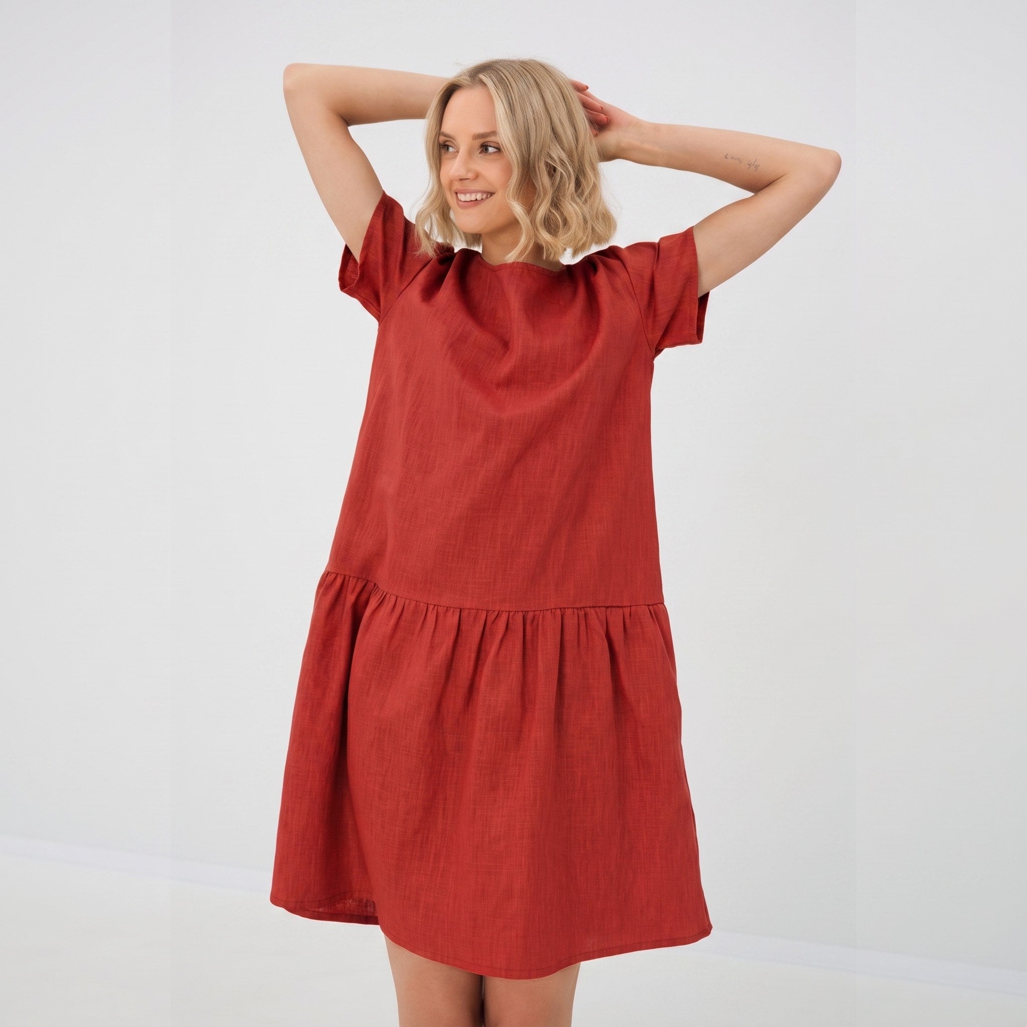 A woman modeling a rust-red short-sleeve linen dress with a tiered waist, standing against a plain white studio background.