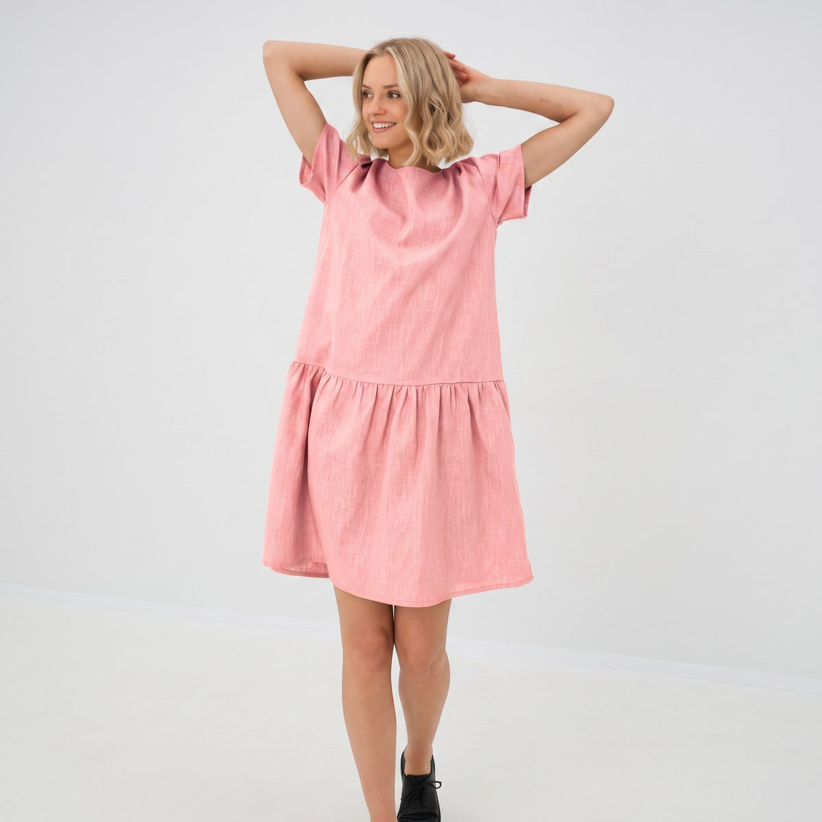 A woman modeling a short-sleeve pink tiered dress with a high round neckline, photographed against a clean white studio background.