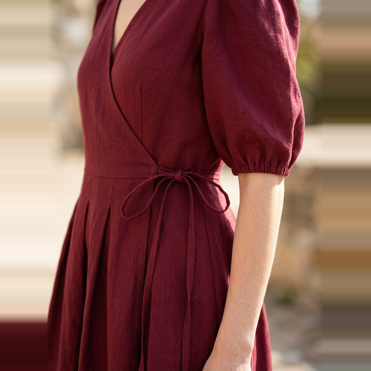 A close-up of a woman wearing a burgundy wrap-style dress with puff sleeves and a tie waist, photographed outdoors against a softly blurred background.