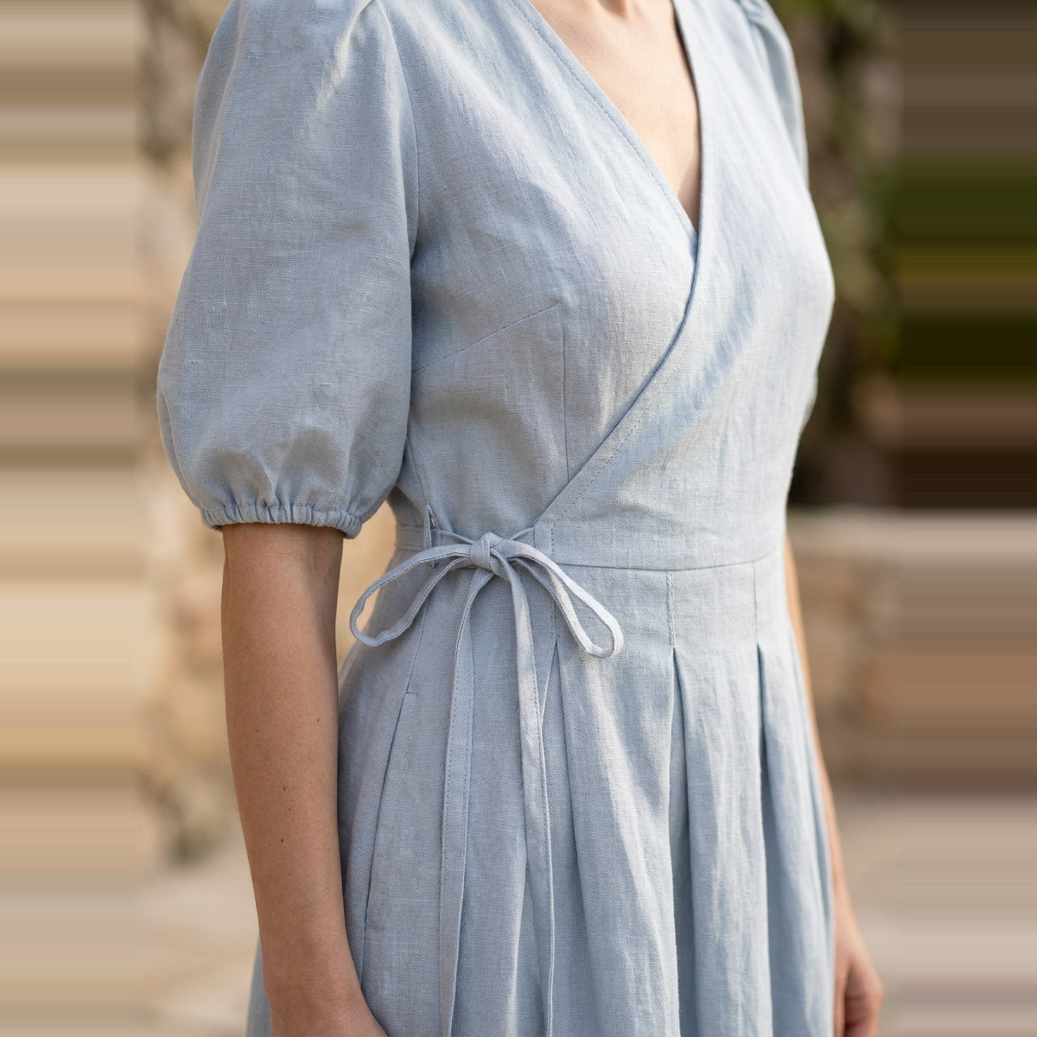 A woman wearing a light blue linen wrap dress with puff sleeves and a tie waist, shown from the chest down against a softly blurred outdoor background.