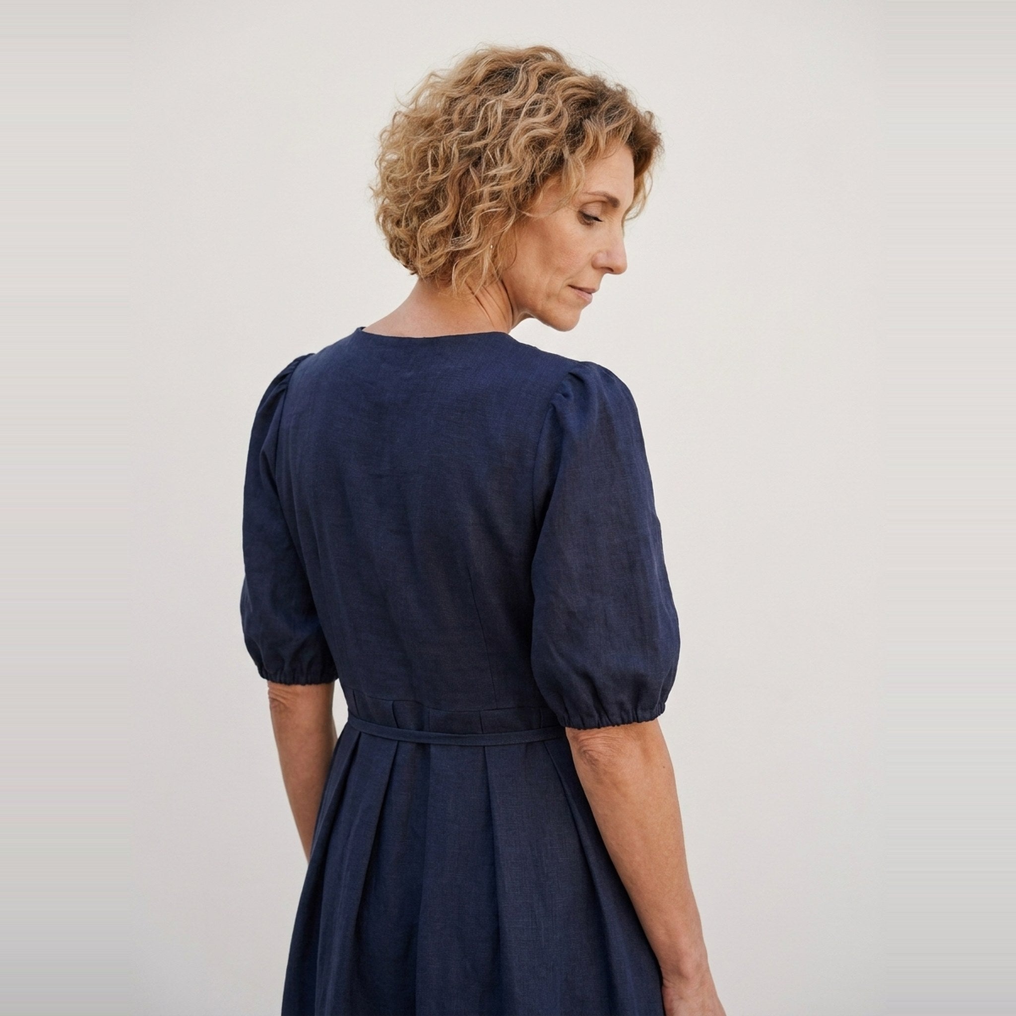A woman with short curly blonde hair is shown from the back wearing a navy blue dress with puff sleeves and a tie waist, photographed against a plain light gray studio background.