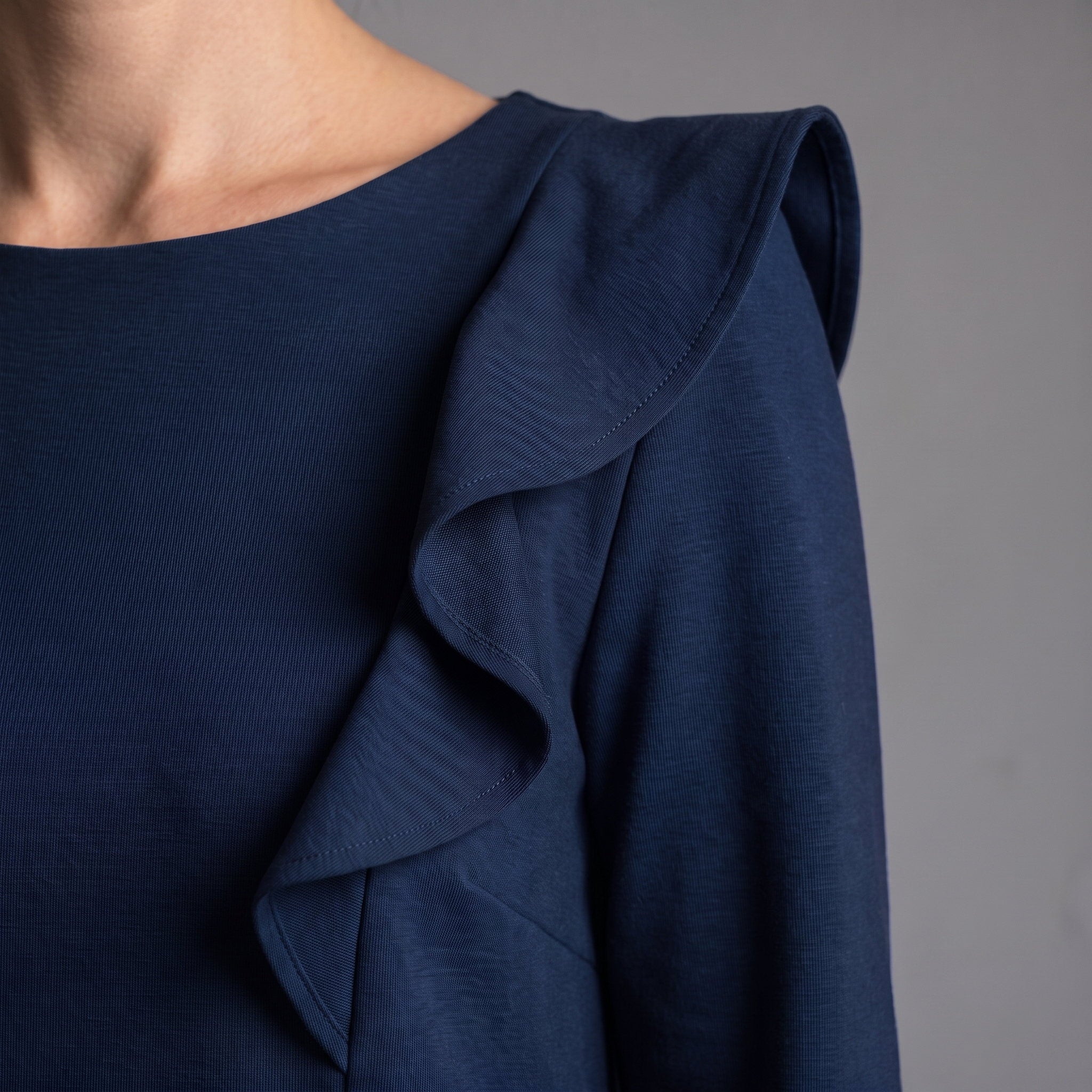 Close-up of a navy blue top with a ruffled shoulder detail, worn by a model against a neutral gray background.