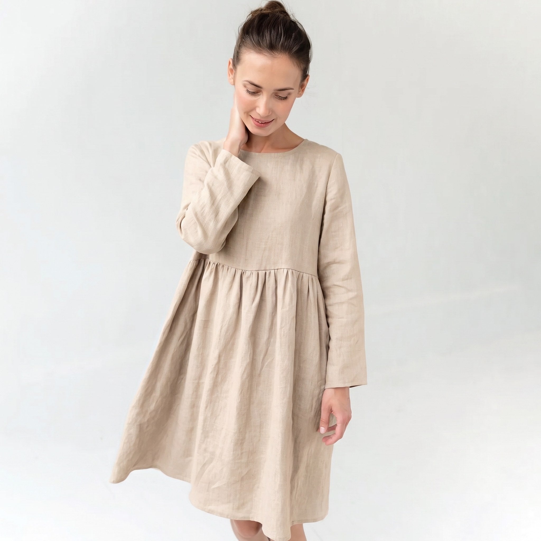 A woman wearing a light beige linen long-sleeve dress with a gathered waist, standing against a plain light gray background.