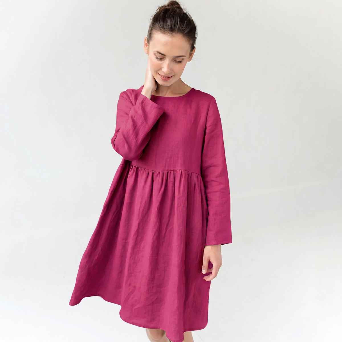 A woman wearing a long-sleeved, tiered linen dress in a deep magenta pink, standing against a plain light gray background.
