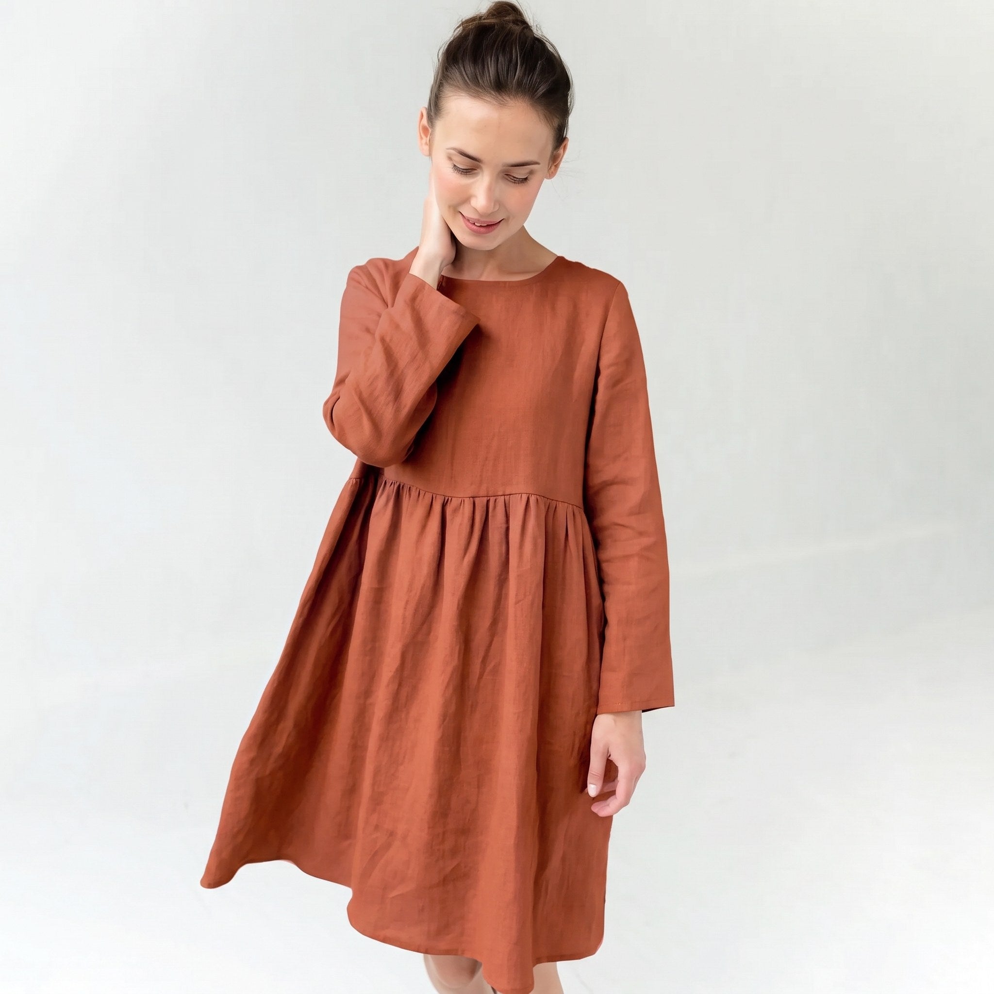 A woman wearing a rust-colored linen dress with long sleeves and a gathered waist, standing against a plain light gray background.