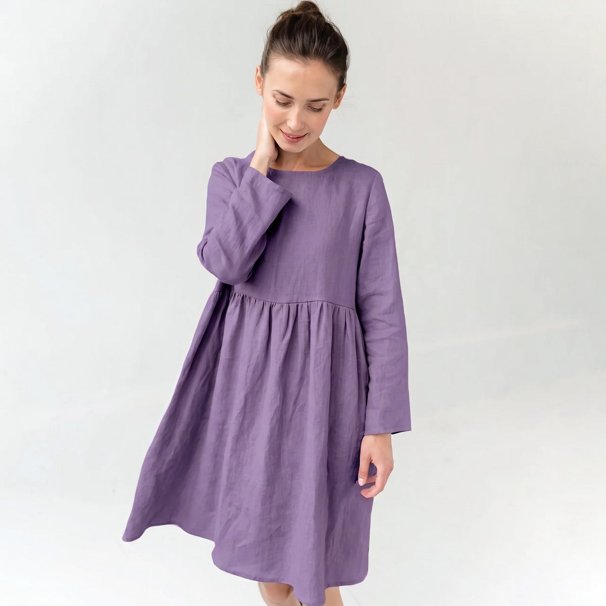 A woman wearing a long-sleeved, knee-length purple dress with a gathered waist, standing against a plain light gray studio background.