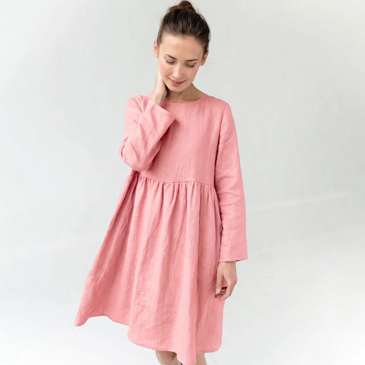 A woman wearing a light pink long-sleeve linen dress with a gathered waist, standing against a plain light gray background.