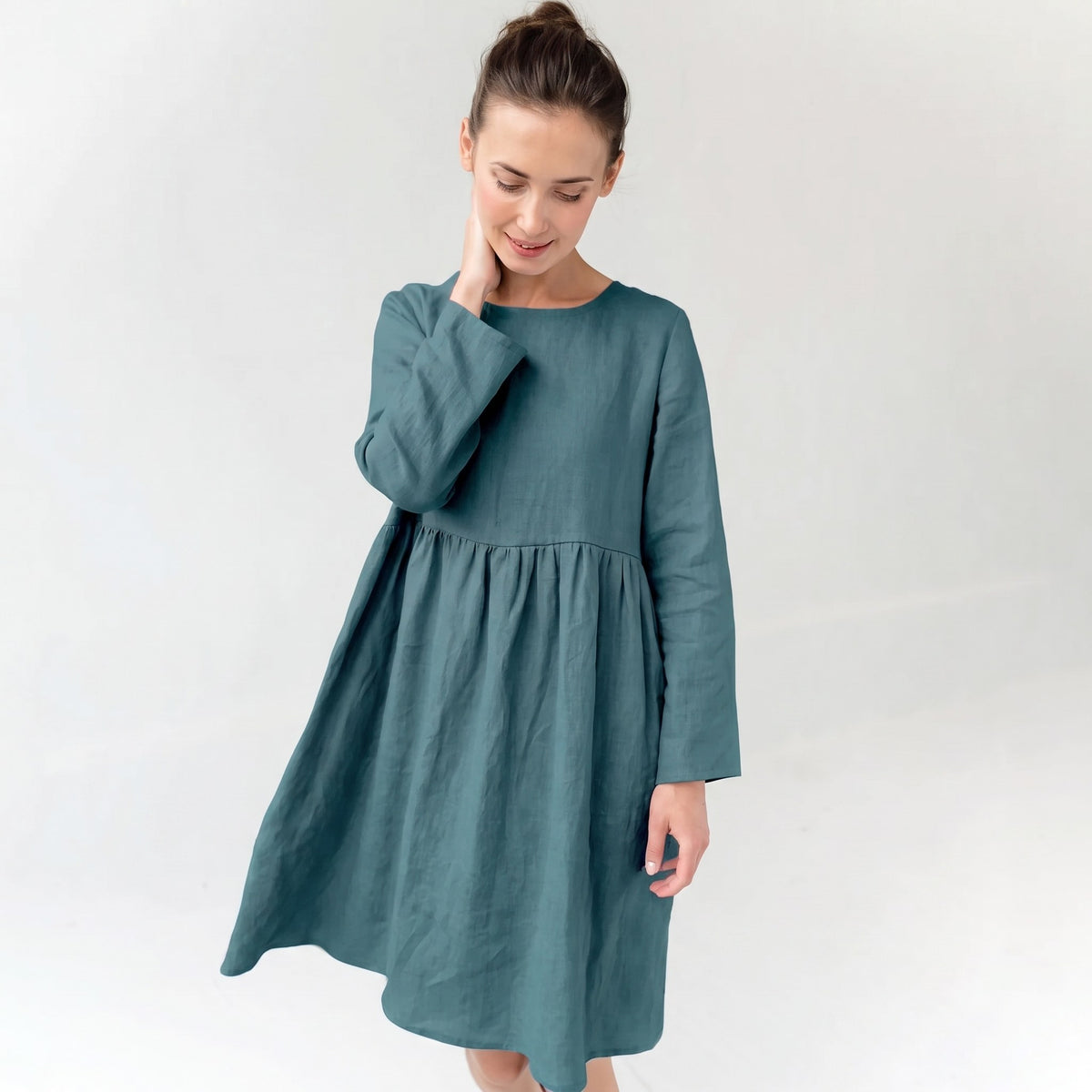 A woman wearing a long-sleeved, knee-length dress in a muted teal blue-green color, standing against a plain light gray studio background.