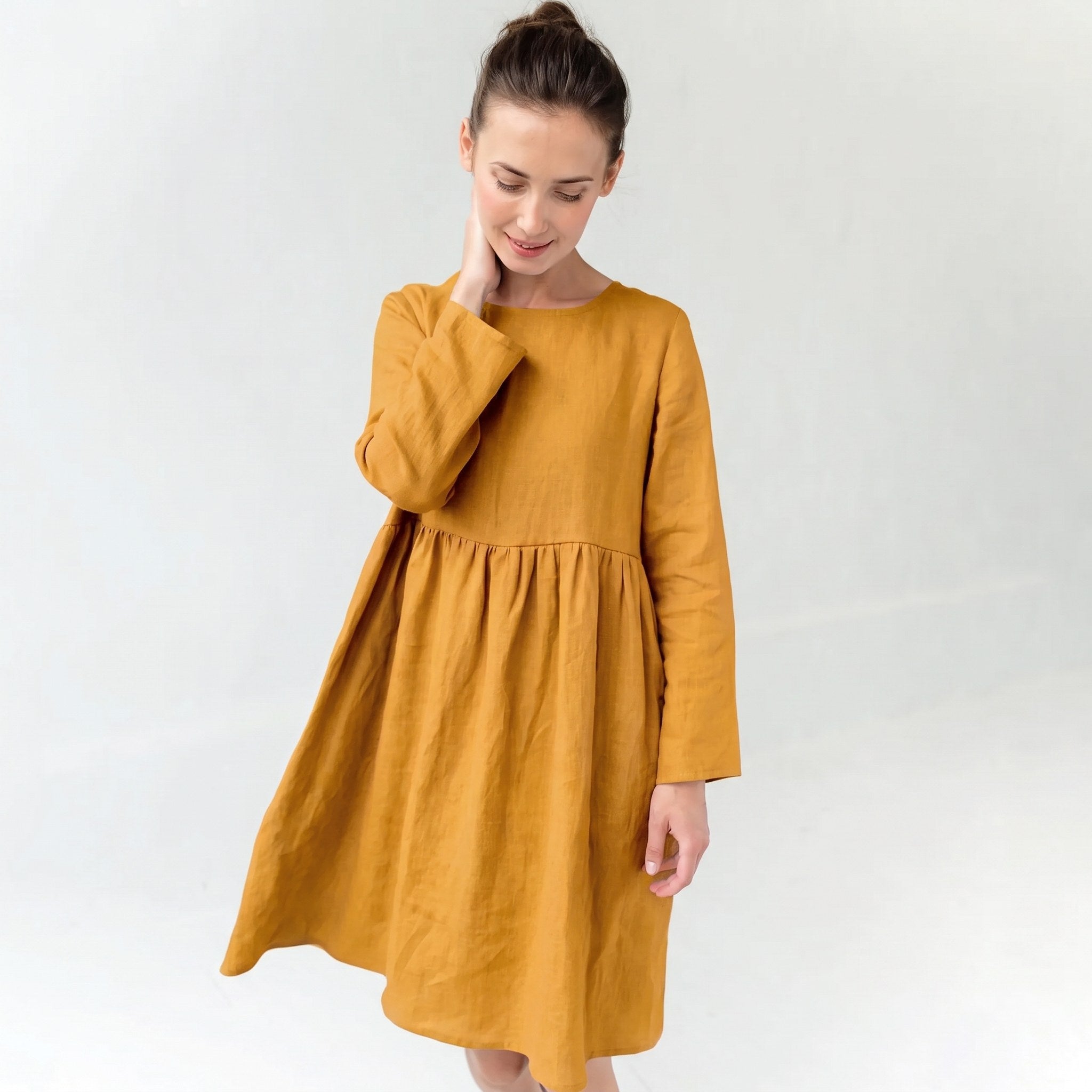 A woman wearing a mustard yellow linen dress with long sleeves and a gathered waist, standing against a plain white studio background.