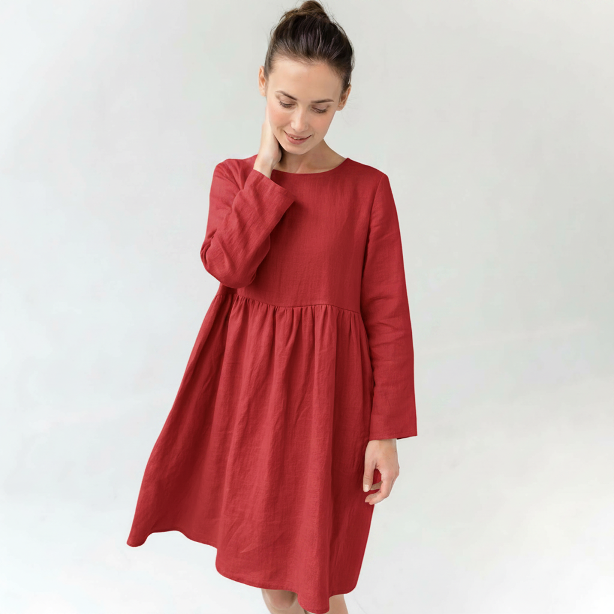 A woman wearing a long-sleeved, red linen dress with a gathered waist, standing against a plain light gray background.