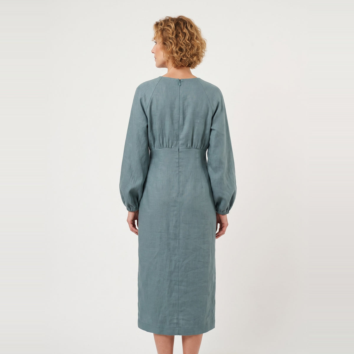 A woman viewed from the back wearing a long-sleeve midi dress in a muted dusty blue-green color, set against a plain off-white studio background.