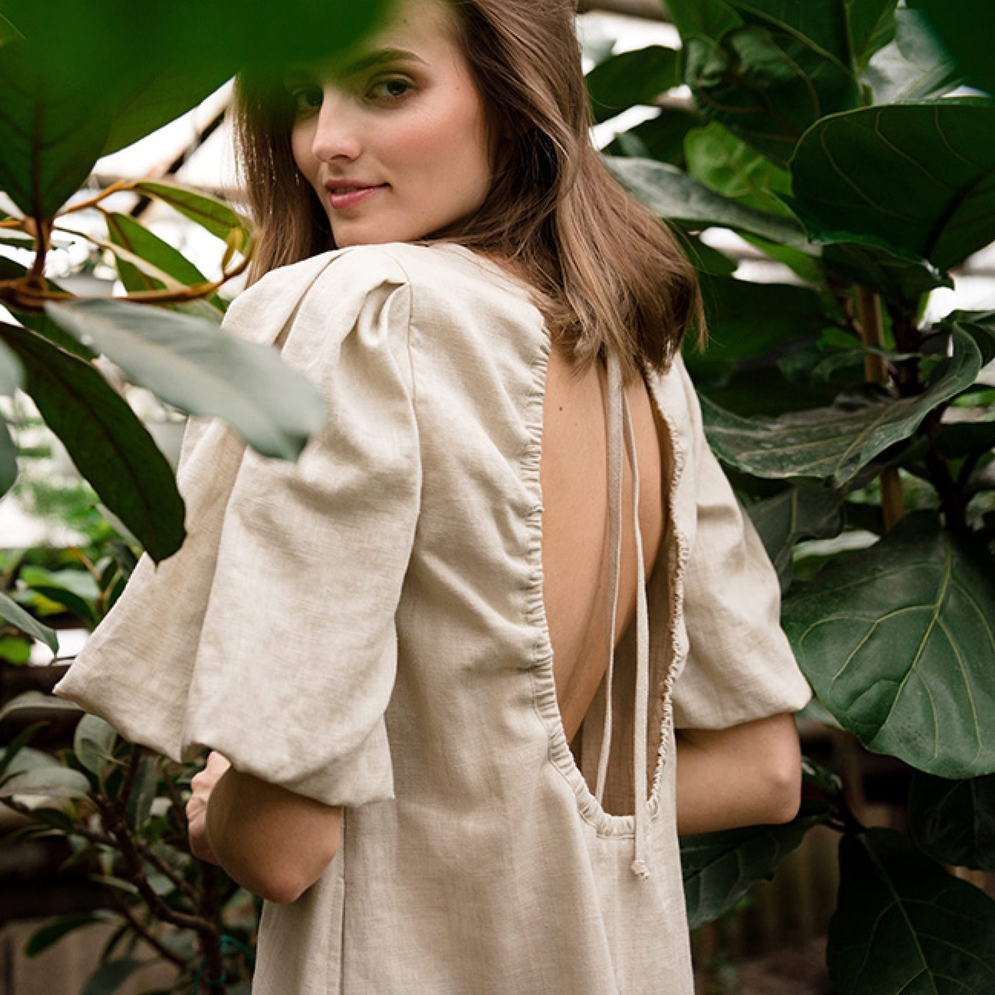 A woman wearing a light beige linen dress with a dramatic open back and puff sleeves, photographed in a lush green garden setting.