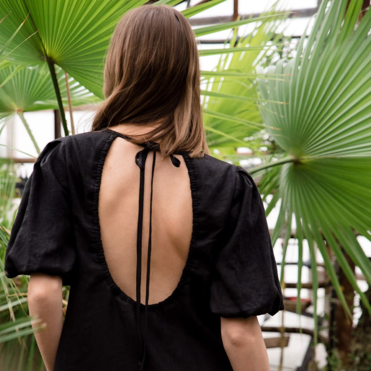 A woman viewed from behind wearing a black short-sleeve top with a large open back tied with thin straps, standing in front of large green palm leaves.