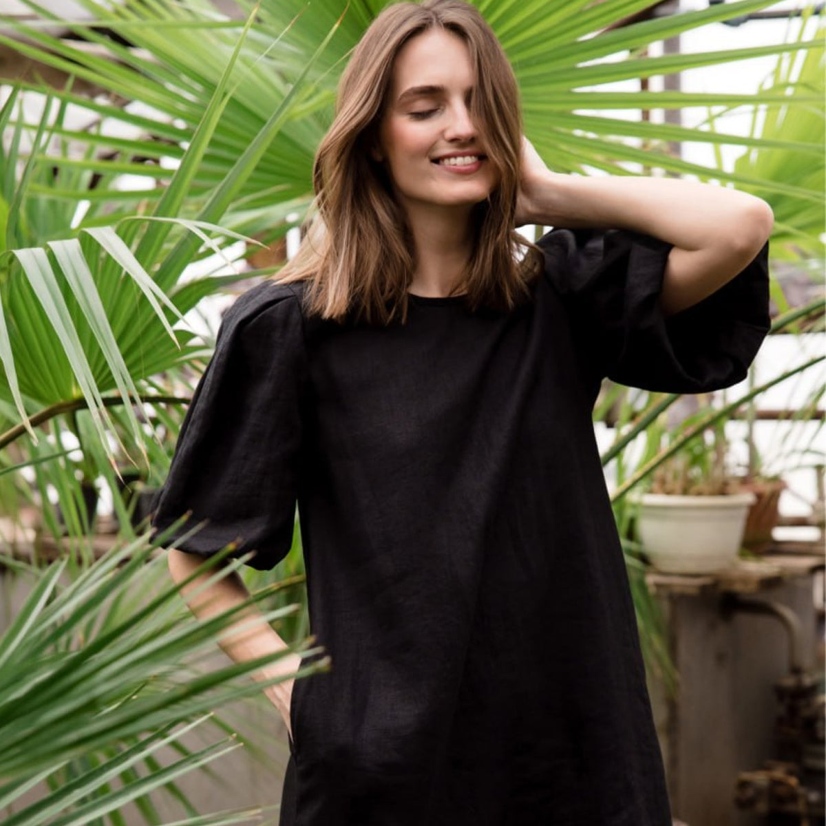 A woman wearing a loose black short-sleeve dress with voluminous puff sleeves, standing in front of lush green palm leaves.