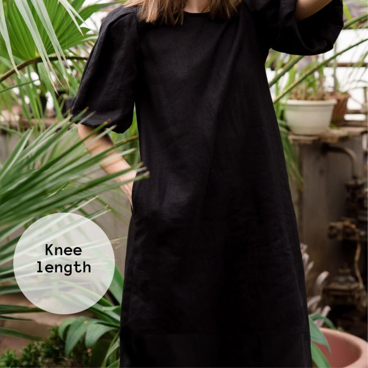 A person wearing a loose black knee-length dress or tunic with short puff sleeves, standing among green plants. A translucent circular label on the left reads 'Knee length'.