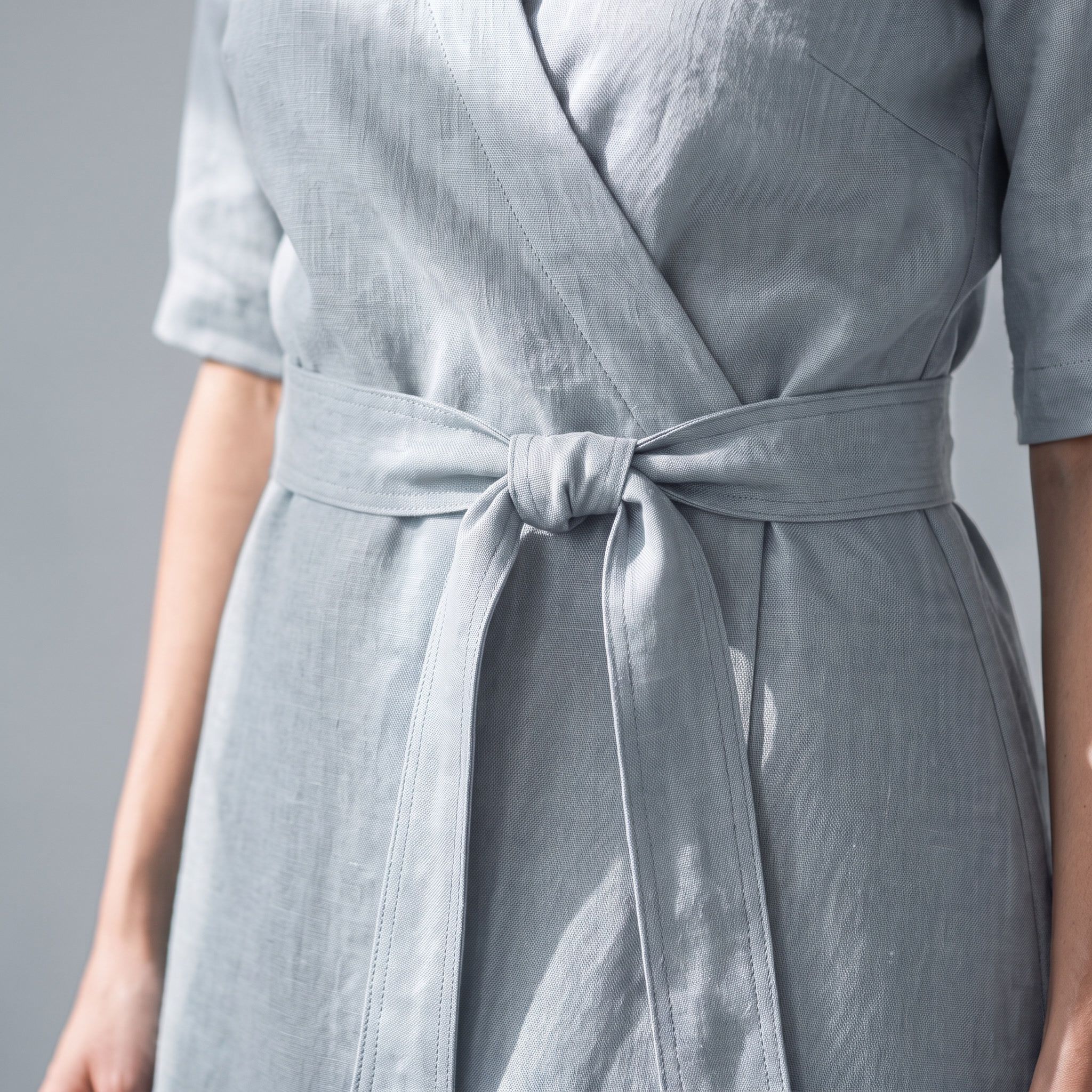 A close-up of a person wearing a light gray wrap-style dress with a matching waist tie, shown against a plain gray background.