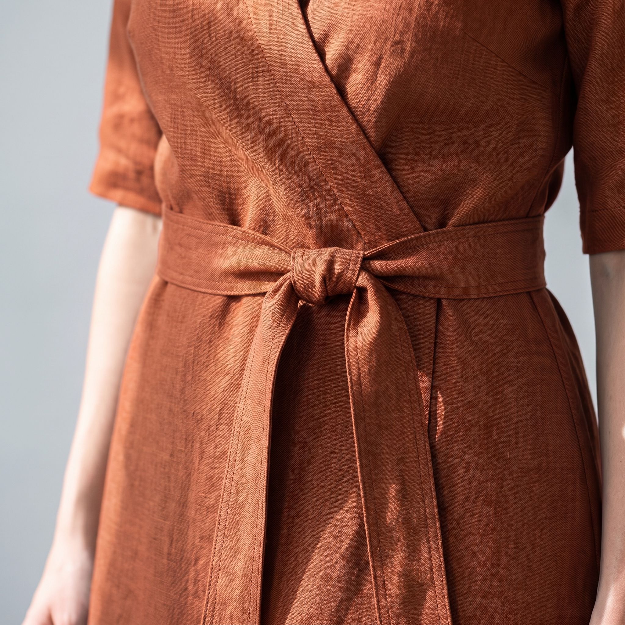A close-up of a terracotta brown linen wrap dress with a matching tie belt, worn by a model against a neutral gray background.