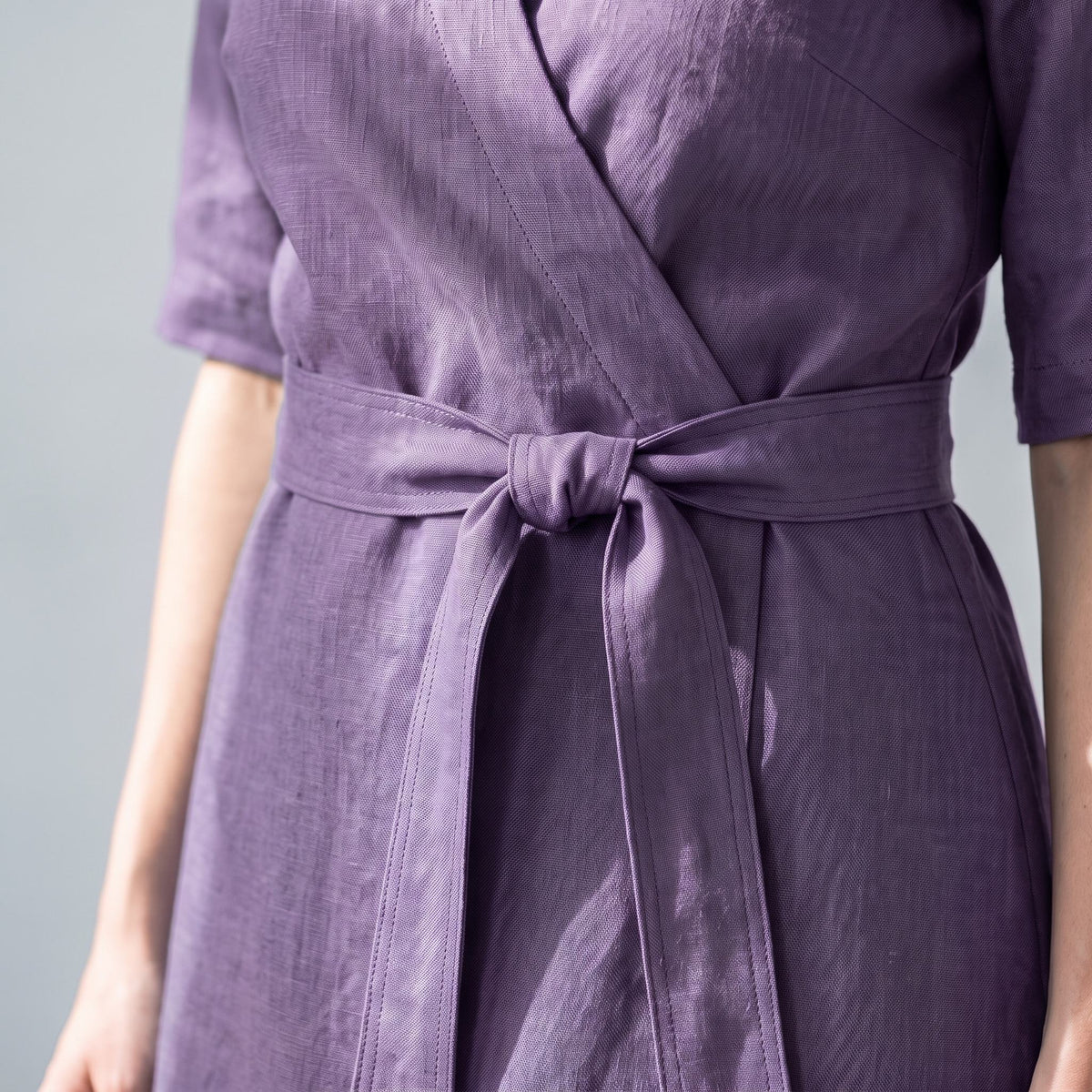 A close-up of a person wearing a lavender-purple linen wrap dress with a matching waist tie, set against a neutral gray background.