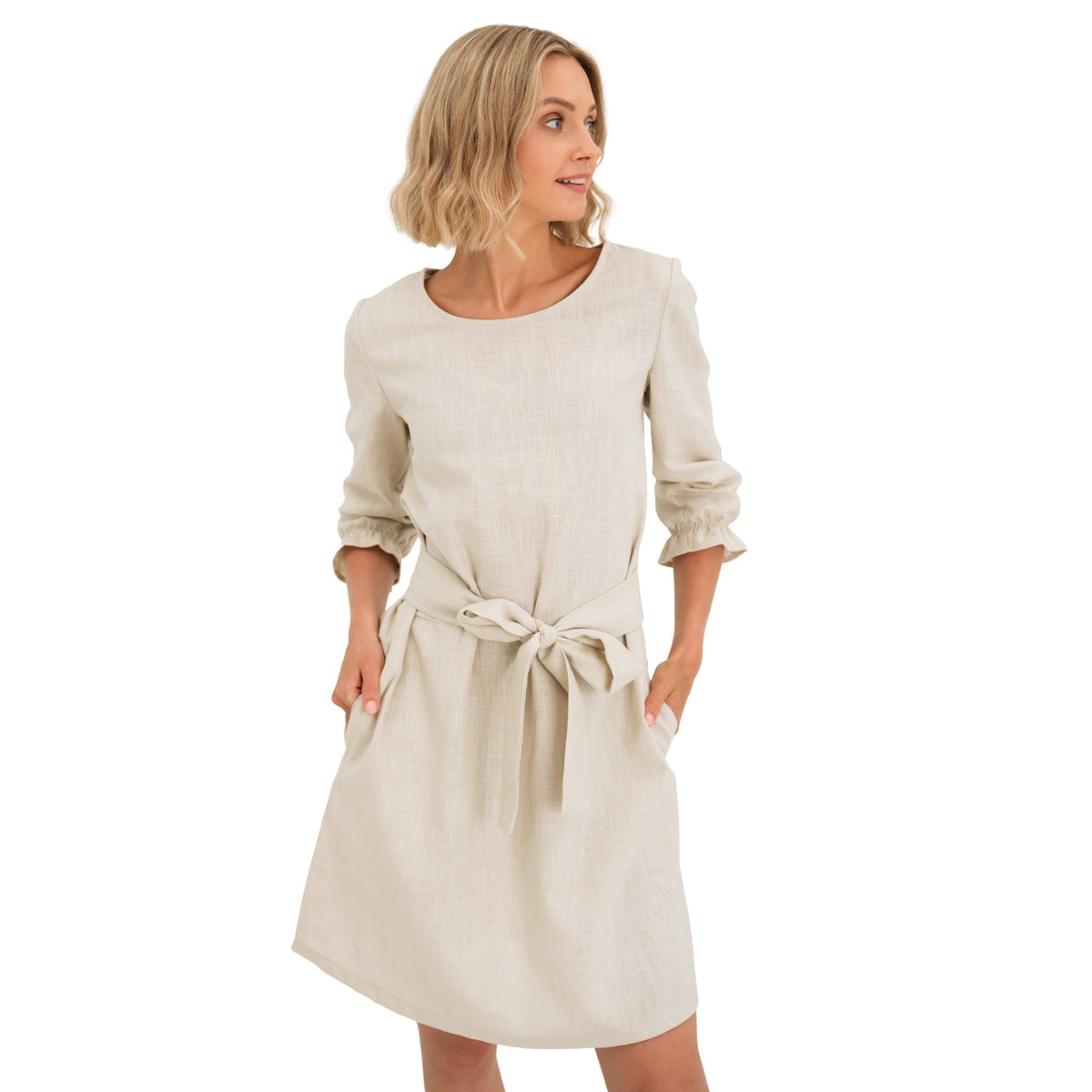 A woman modeling a light beige linen dress with three-quarter sleeves, a matching fabric tie belt at the waist, and side pockets, set against a plain white background.