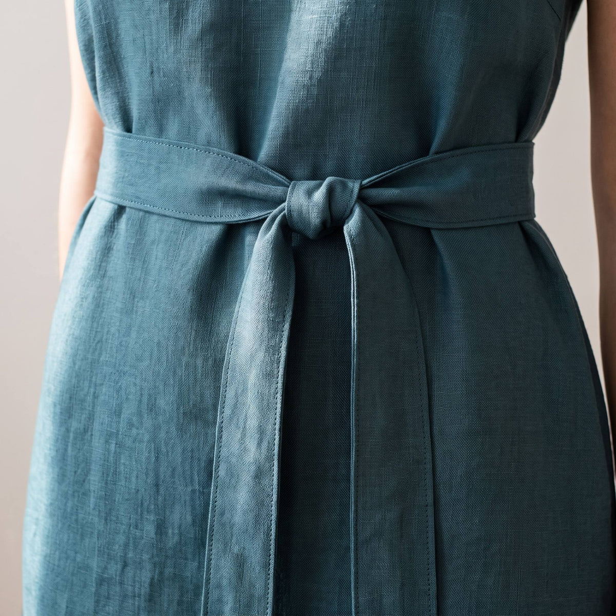 A close-up of a sleeveless blue-green dress with a matching fabric tie belt knotted at the waist, shown on a model against a soft neutral background.