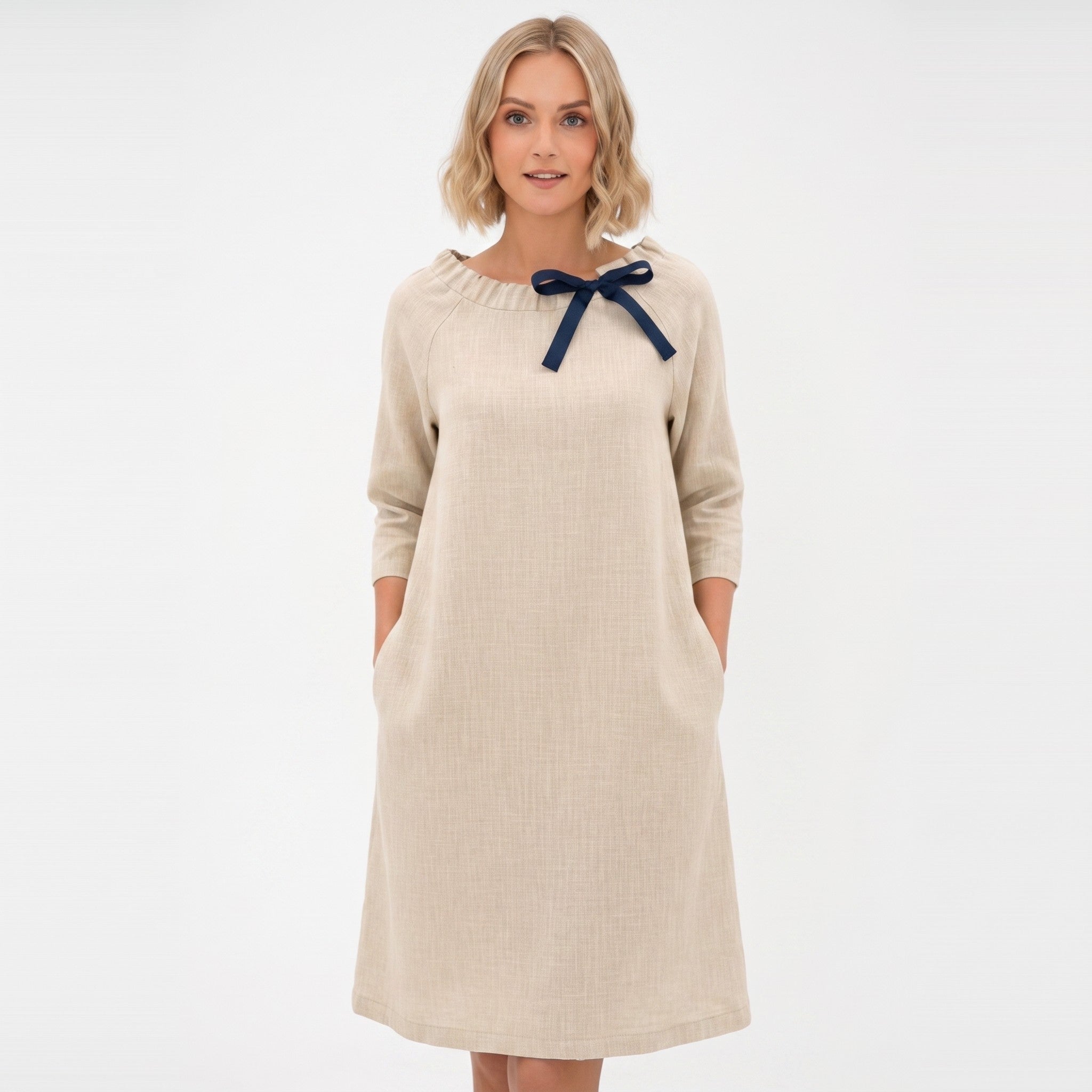 A woman modeling a beige long-sleeve dress with a navy blue bow detail at the neckline, set against a plain white background.