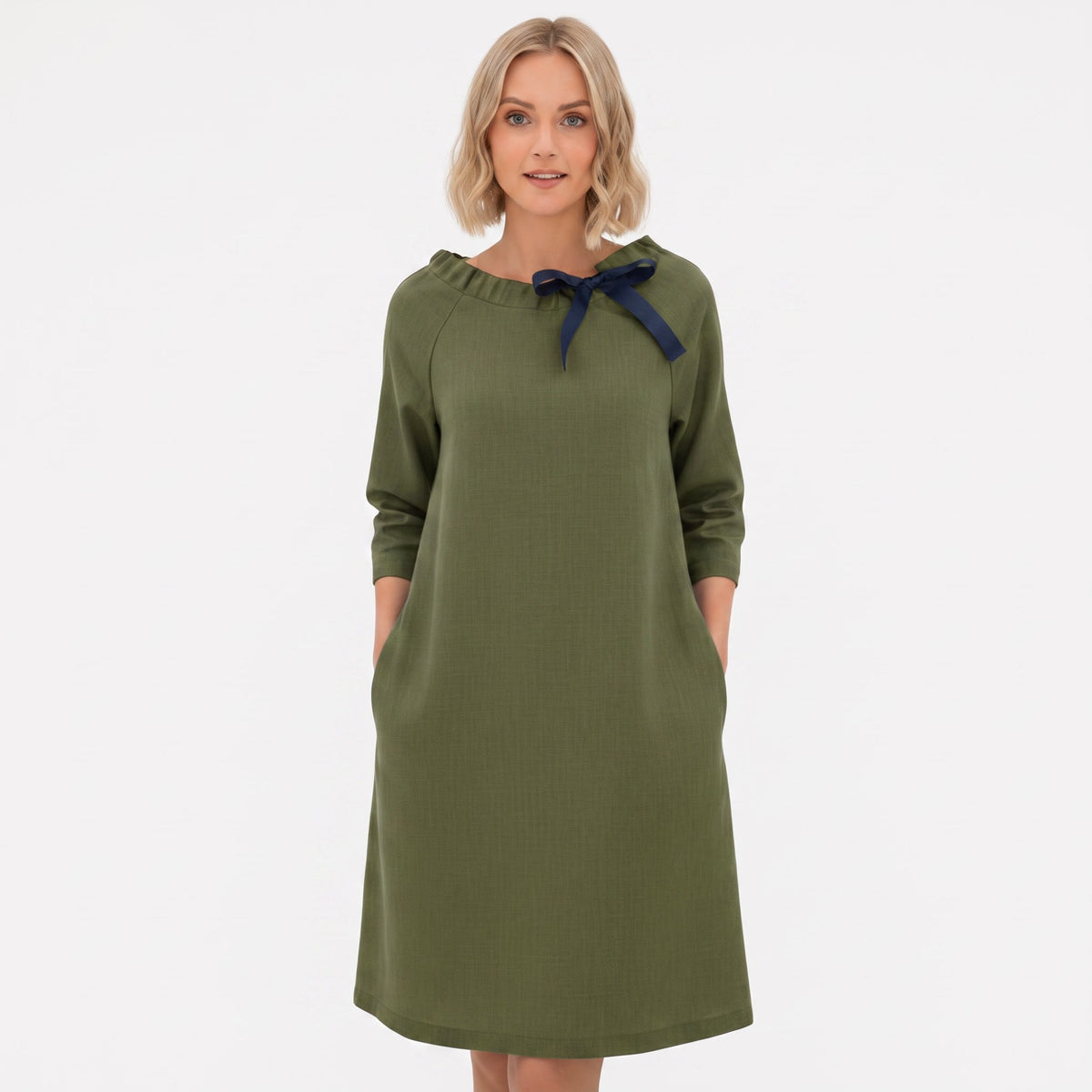 A woman modeling an olive green long-sleeve dress with a navy blue bow detail at the neckline, set against a plain white background.
