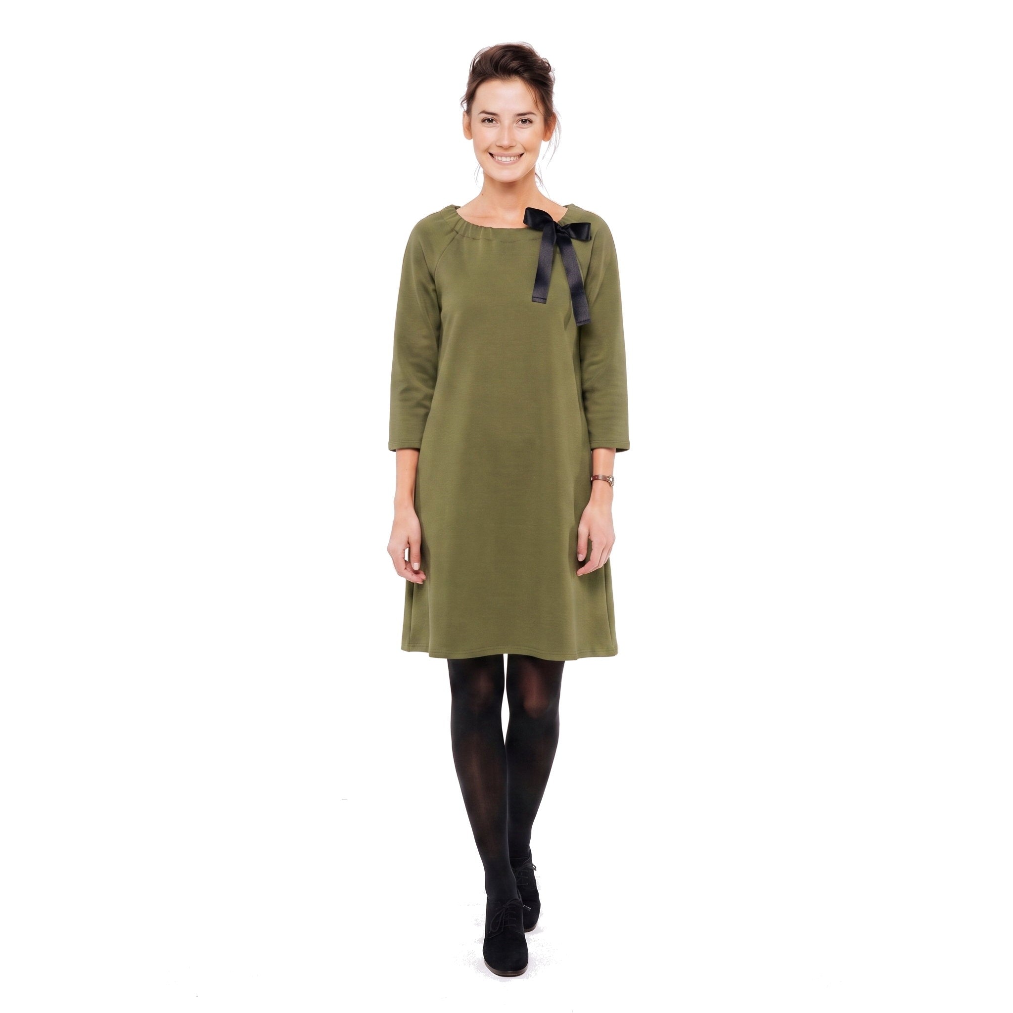 A woman modeling an olive green dress with three-quarter sleeves and a black ribbon bow at the neckline, paired with black tights and black ankle boots, against a plain white background.
