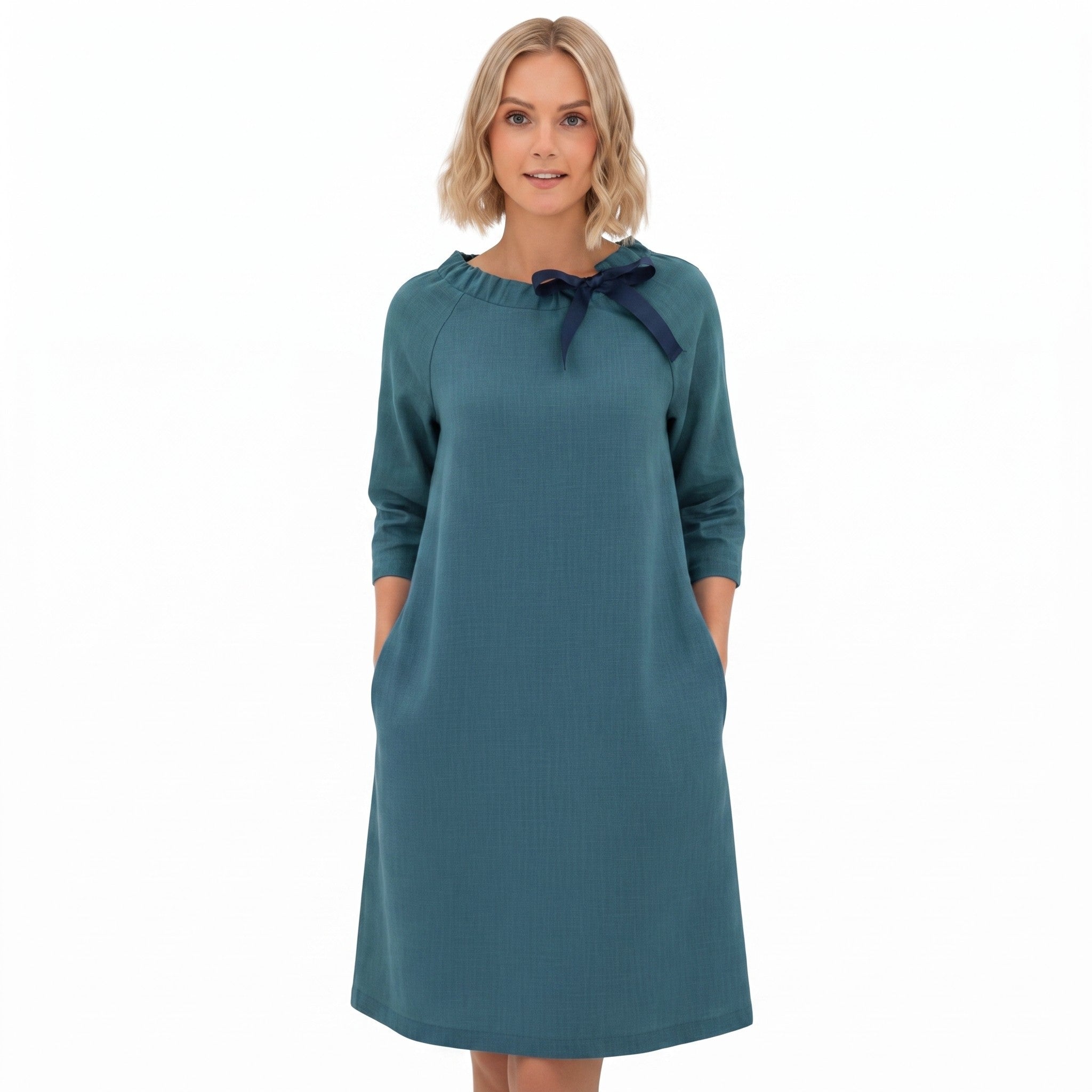 A woman modeling a teal blue long-sleeve dress with a navy blue bow detail at the neckline, set against a plain white background.