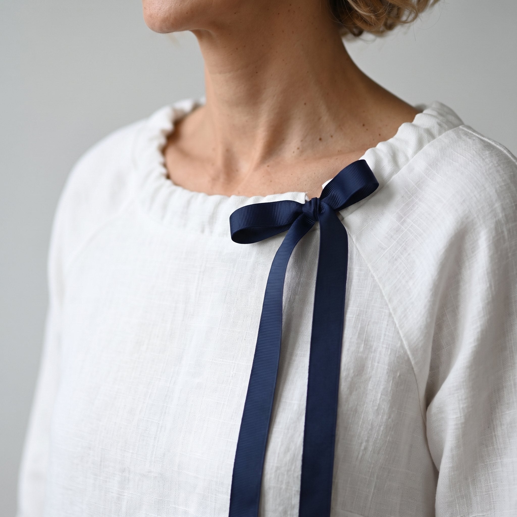 A close-up of a person wearing a white textured linen top with a dark navy ribbon tied in a bow at the ruffled neckline.