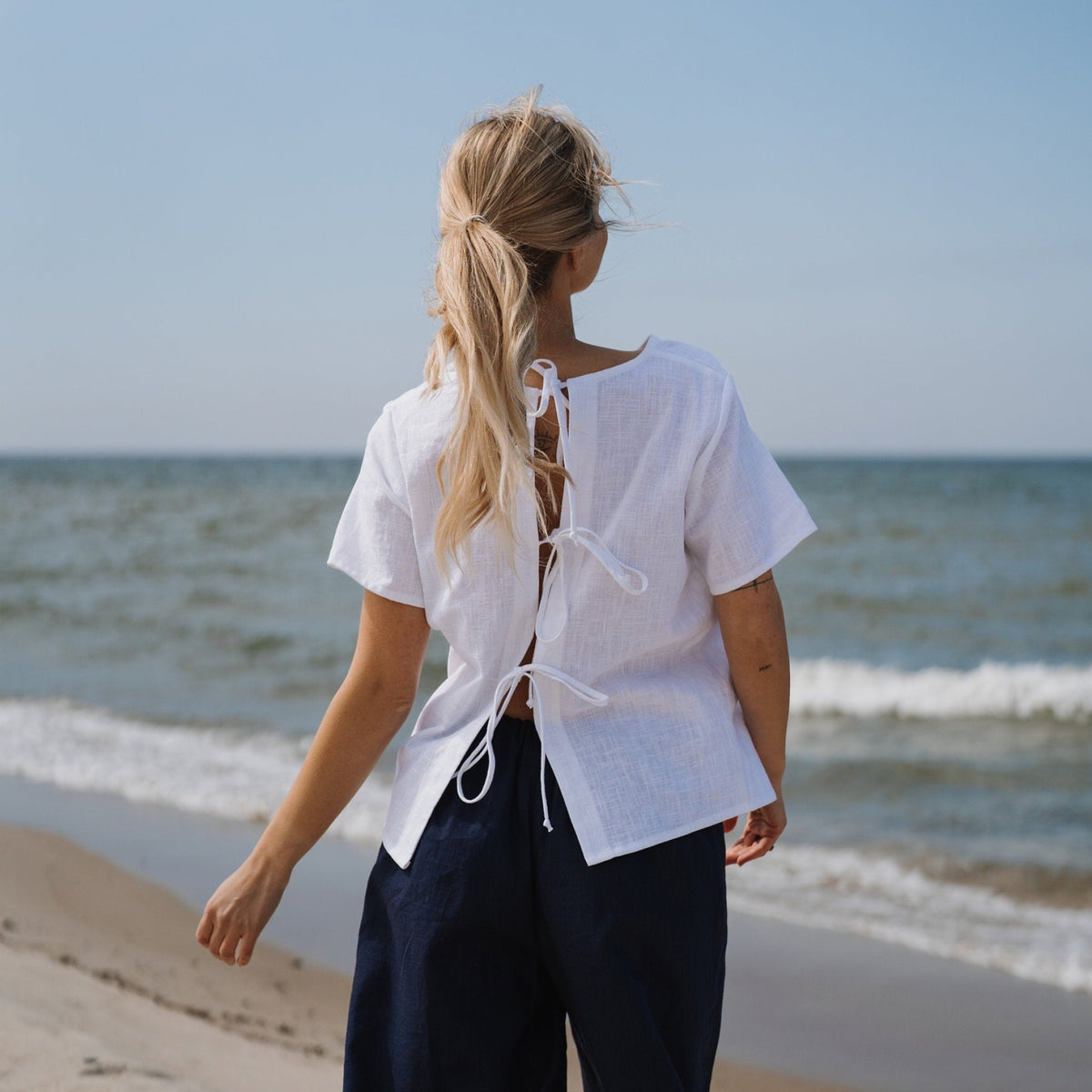 A woman stands on a sandy beach facing the ocean, wearing a white short-sleeve linen blouse with lace-up back details and dark navy pants.