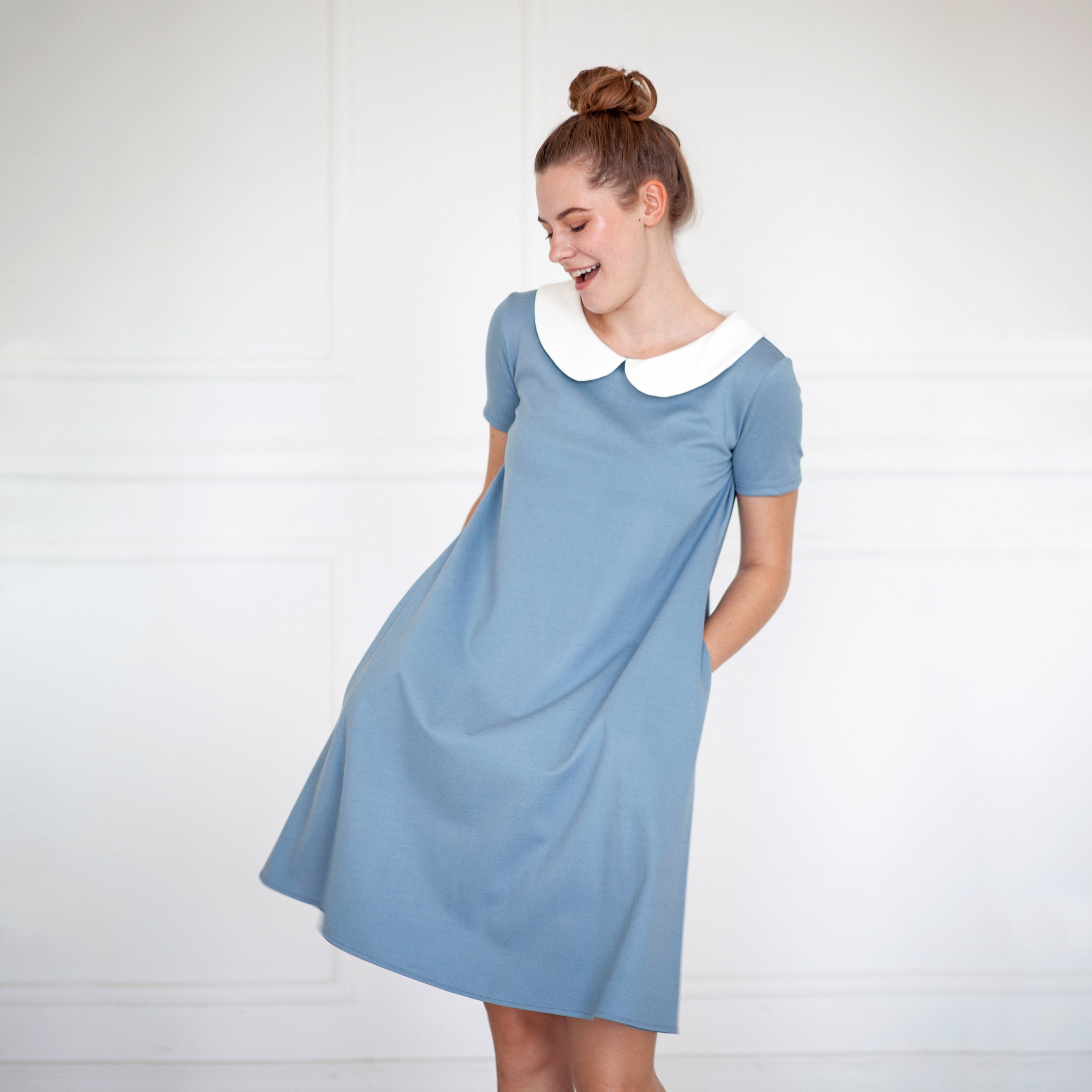 A woman is posing in a deep blue knee-length dress with a white rounded collar and short sleeves. The dress has a pockets on the sides, and the fabric appears to be viscose.