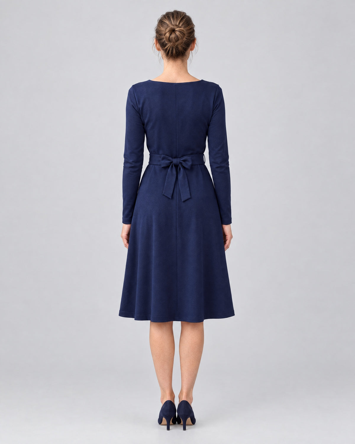 A woman wearing a navy blue long-sleeve V-neck Punto Milano dress with a tie belt bow, back view, full length. #Color_Deep blue