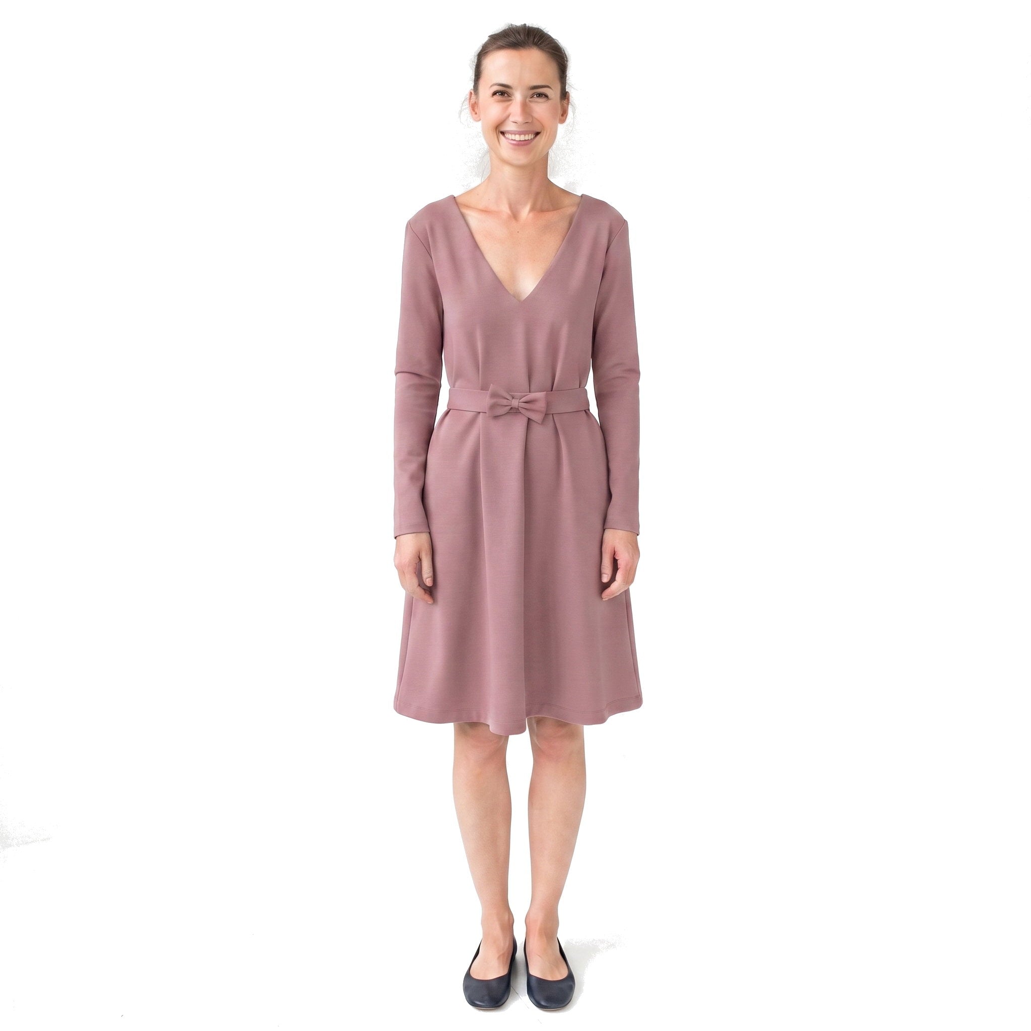 A woman modeling a dusty rose long-sleeve V-neck dress with a matching fabric tie belt, paired with black flats, against a plain white background.