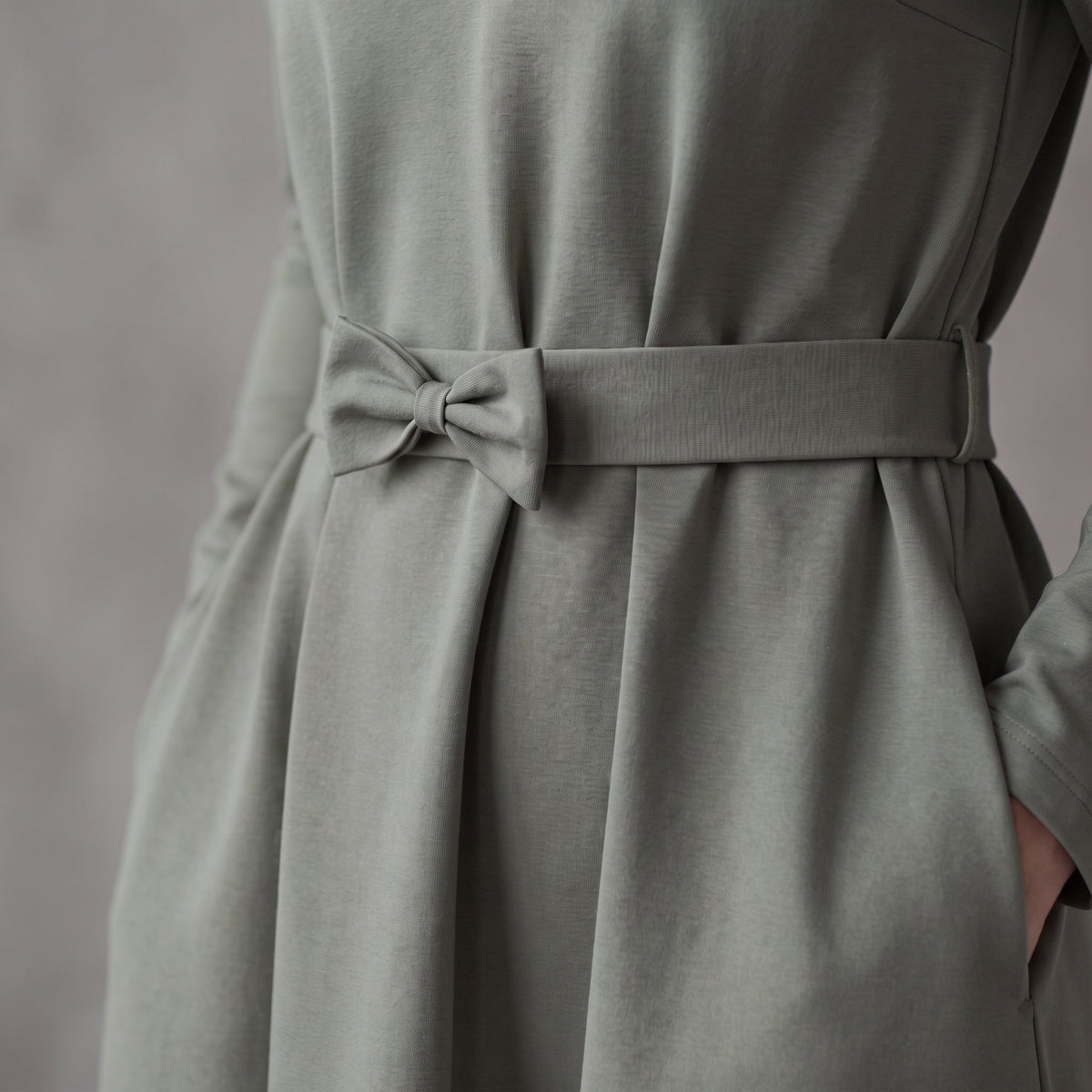 A sage green dress with a matching fabric tie belt at the waist, showing the draped skirt and side pocket detail.