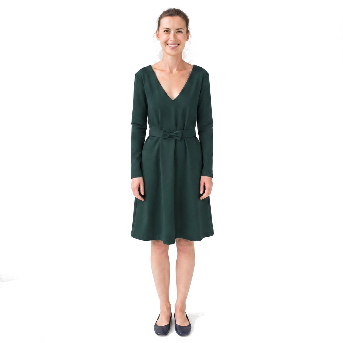 A woman modeling a dark green long-sleeve V-neck dress with a matching waist tie, paired with dark navy flats, against a plain white background.