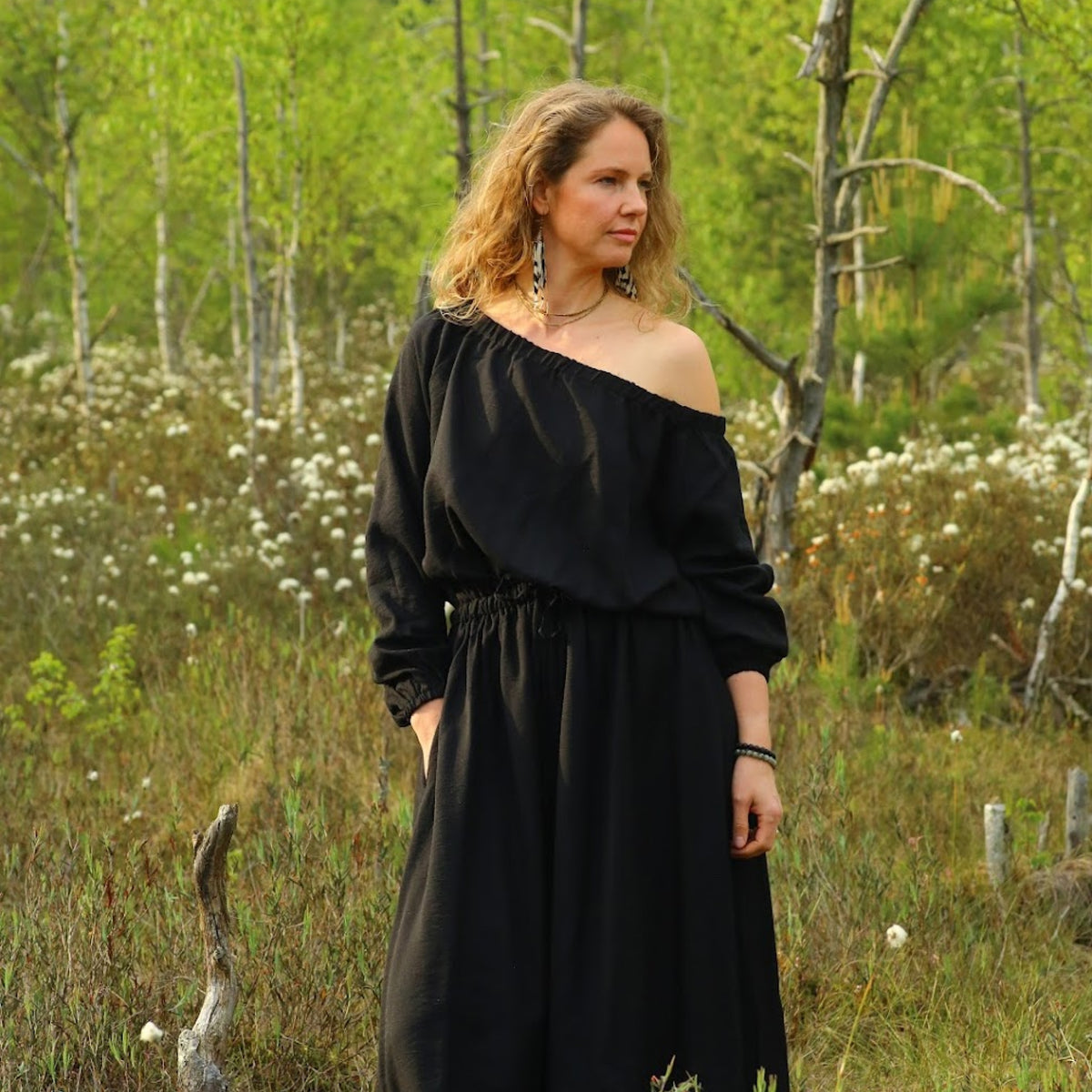 A woman wearing a black off-the-shoulder maxi dress stands in a grassy, wooded outdoor setting with birch trees and small white flowers.