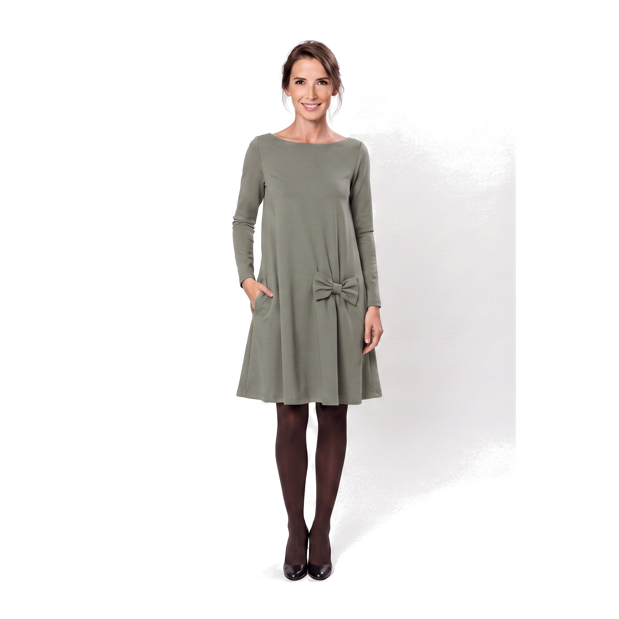 A woman modeling a olive green long-sleeve A-line dress with a boat neckline, side pockets, and a bow detail at the waist.