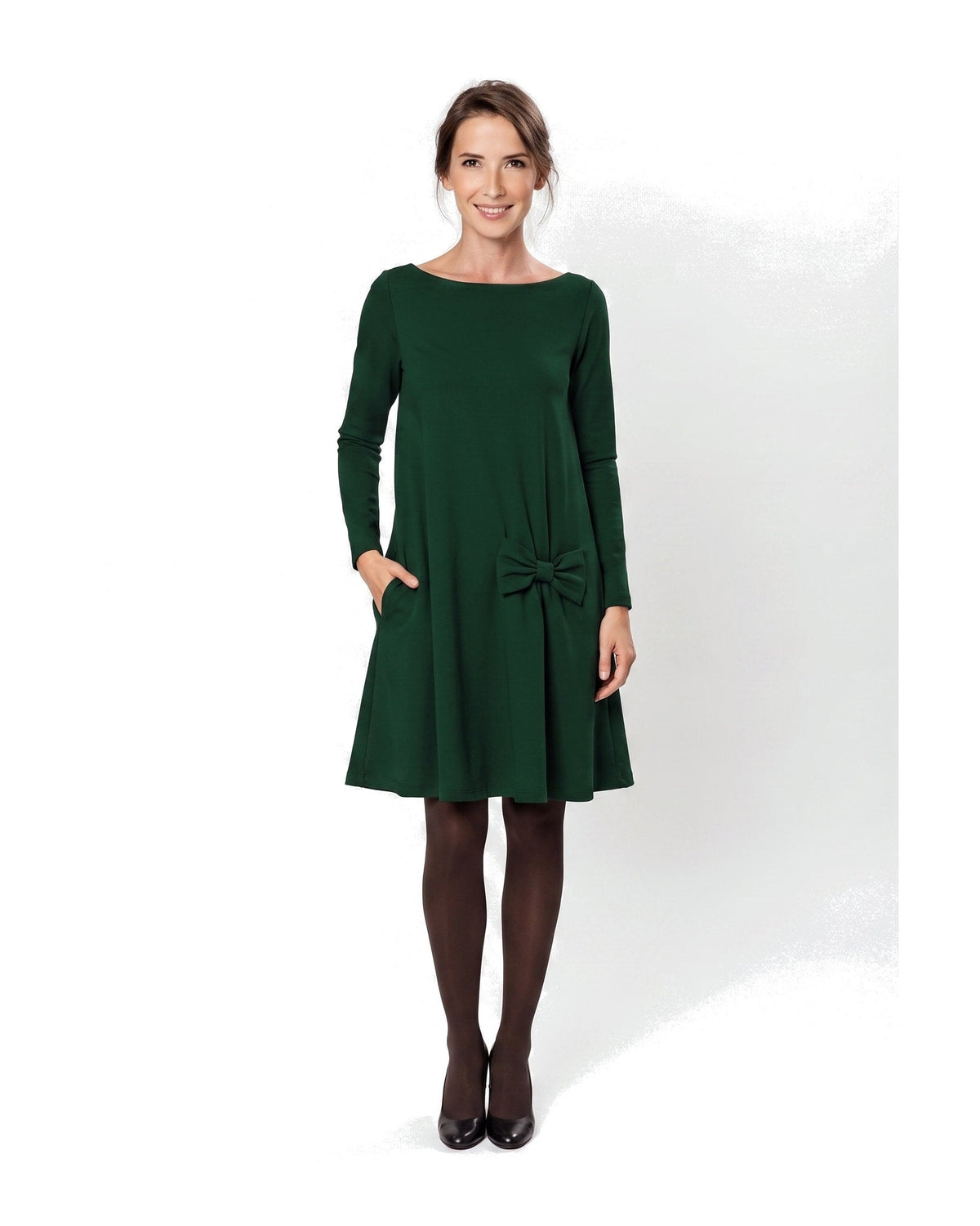 A woman modeling a dark green long-sleeve A-line dress with a boat neckline, side pockets, and a bow detail at the waist. #Color_Green