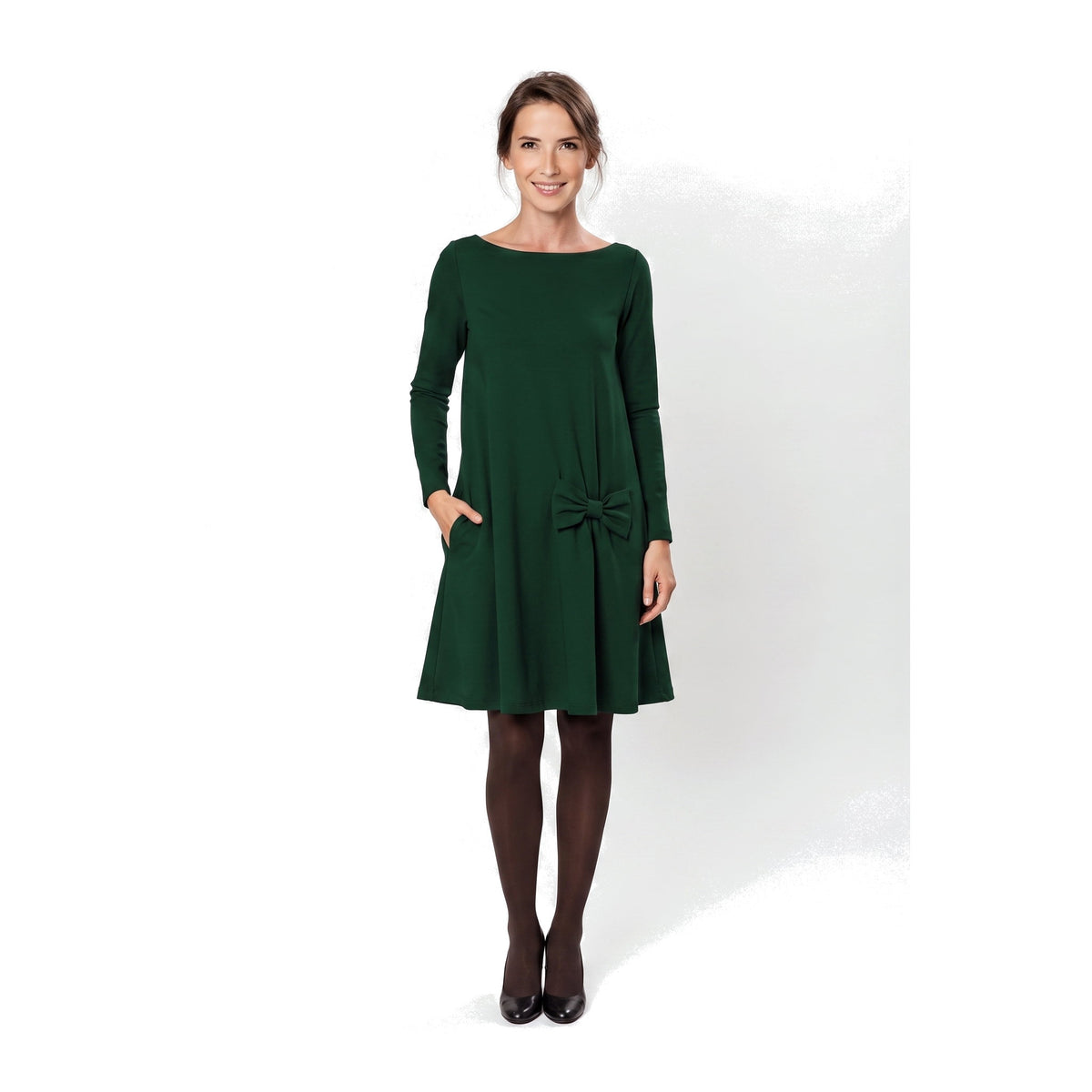 A woman modeling a dark green long-sleeve A-line dress with a boat neckline, side pockets, and a bow detail at the waist.
